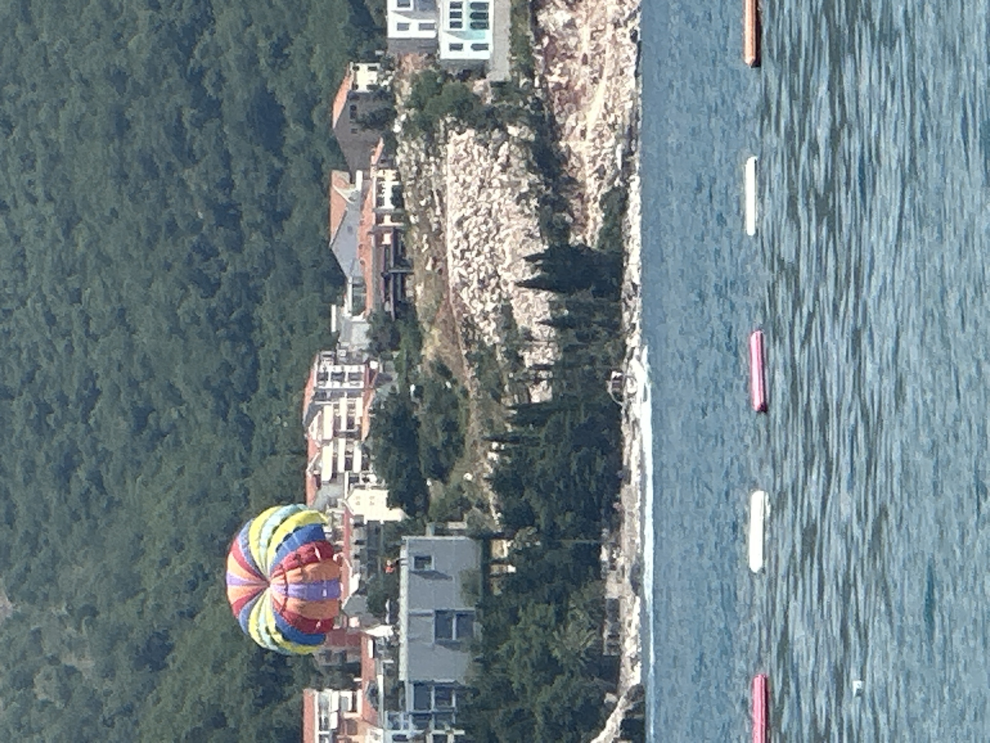 The image depicts a coastal scene in Bečići, Montenegro. The foreground is dominated by the deep blue, rippling water of the sea, marked by several buoys that appear to be for swimming or a water sport. One buoy is a distinct pink, another is white, and a third, further away, is also pink. The water's surface reflects the bright sunlight, indicating a clear day.
Adjacent to the water, a shoreline is visible, with a rocky embankment dotted with trees and some buildings. This suggests a developed coastal area, likely a resort or town. The buildings are varied in style, some appearing to be modern apartments or hotels with white facades and balconies, while others have terracotta roofs, hinting at a Mediterranean architectural influence.
Further inland, a dense, dark green forest covers a steep hillside, providing a stark contrast to the built-up area and the sea. The forest suggests a mountainous or hilly terrain behind the coast.
Dominating the mid-ground and left side of the image is a colorful parasail, a large, multi-colored parachute, suspended in the air. Its vibrant segments of red, yellow, blue, and purple suggest it is in use, likely for a recreational activity. The parasail is positioned over the water, indicating a parasailing experience is taking place. The perspective suggests the photo is taken from a higher vantage point, looking down and across the scene. The overall impression is one of a sunny, active vacation spot. No text is visible in the image.