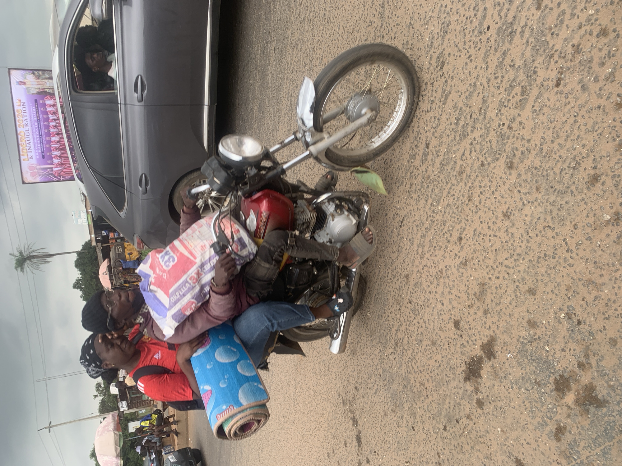 A motorcycle carrying two individuals and a large rolled-up blue mat is moving on a paved road. The person in the front, wearing glasses and a black beanie, is driving the motorcycle. The person behind, wearing a red shirt with black stripes on the sleeves and a black bandana, is holding the blue mat. The motorcycle is red and appears to be a 2014 Bajaj Boxer model. A gray Lexus SUV is driving in the same direction behind them. A billboard promoting "Lidero 2020 Iv & Inauguration" is visible in the background, suggesting the location is Shagamu, Nigeria. The environment appears to be a roadside with some businesses and trees. The sky is overcast.