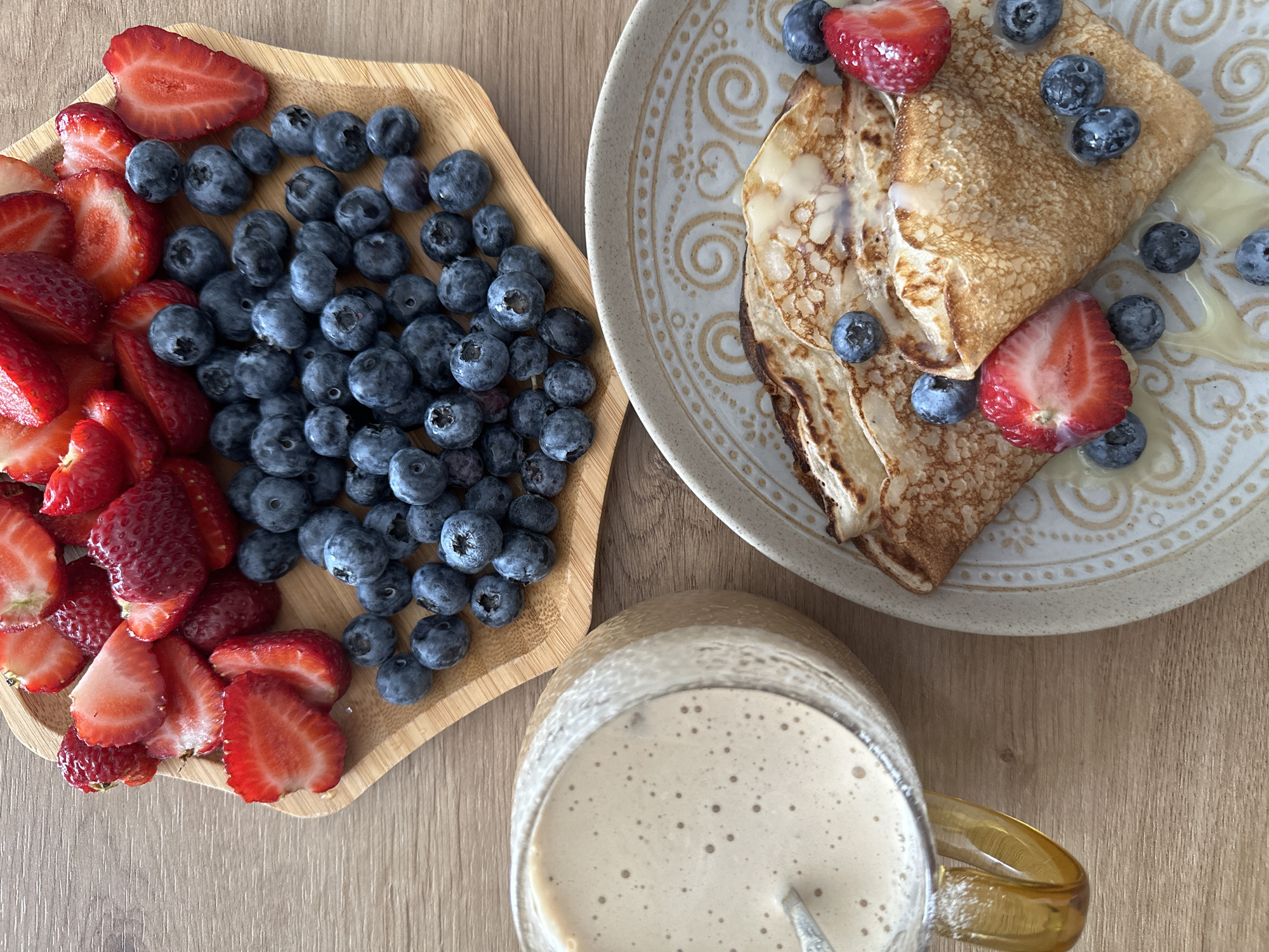 A wooden table with a breakfast spread. On the left, a glass of light brown liquid with a spoon submerged in it. In the center, a hexagonal wooden dish filled with sliced strawberries and blueberries. On the right, a plate with two crepes, garnished with blueberries and sliced strawberries, drizzled with a light-colored syrup. The background is a wooden surface. There is no indication of the city of Fethiye, Turkey.