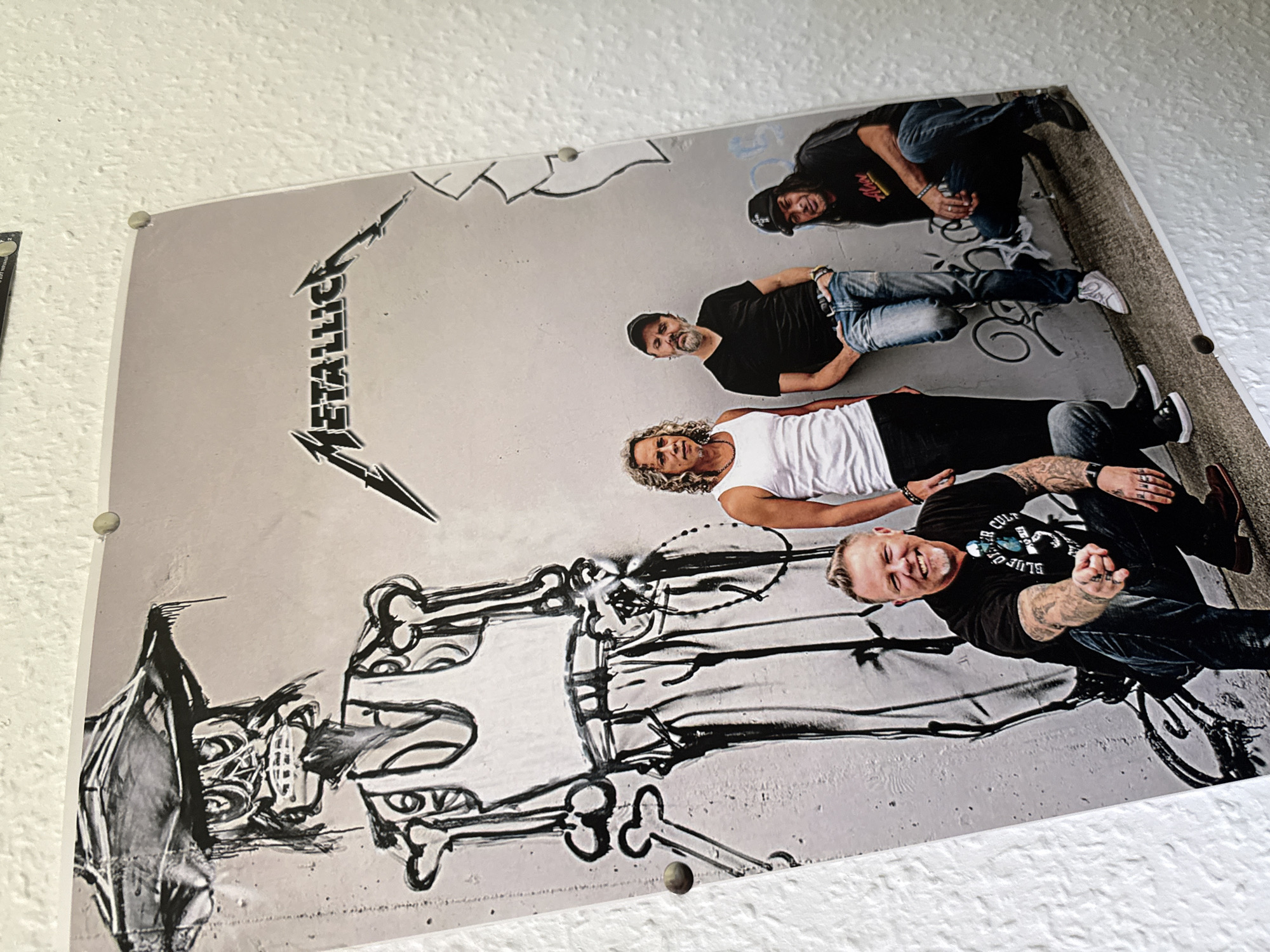 The image shows a poster of the band Metallica on a textured white wall, possibly in the town of Bad Sachsa, Germany, given the context provided. The poster features a large, stylized skeletal figure in a hat and tank top on the left, and the band members on the right. James Hetfield, wearing a black t-shirt with "Blue Öyster Cult" text and blue sunglasses, is in the foreground, crouched and smiling with his fist extended. To his right, Lars Ulrich, in a black t-shirt and jeans, leans against the wall. Kirk Hammett, with long curly hair and a white tank top, stands next to Ulrich. Robert Trujillo, wearing a black t-shirt, jeans, and a black baseball cap, is crouched on the far right. The background appears to be a graffitied wall. The poster is secured to the wall with round fasteners, visible at the top and bottom.