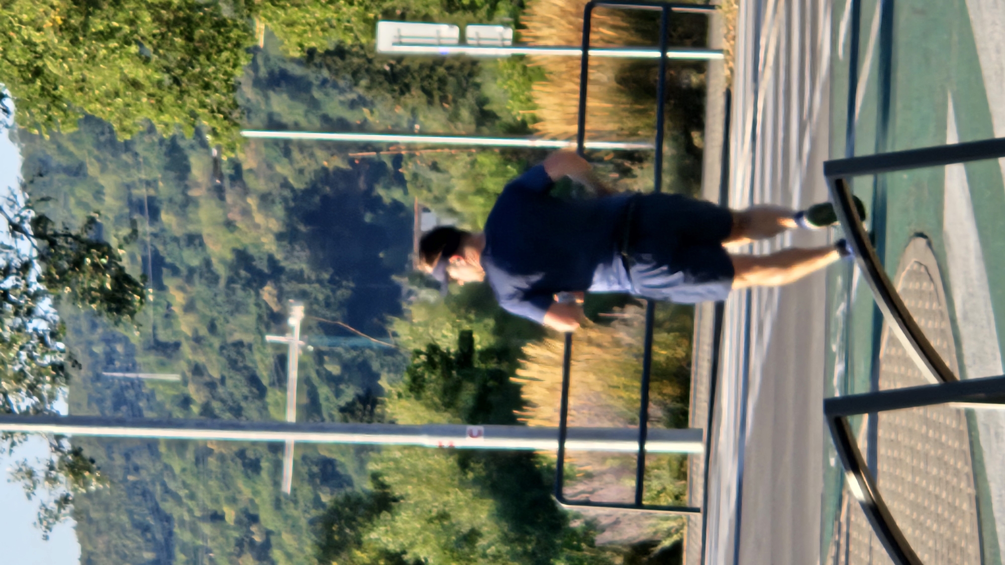 An adult male is running in an outdoor urban setting, facing away from the camera. He is wearing a dark blue t-shirt, dark blue shorts, and a dark visor. He is on a paved surface, possibly a pedestrian path or road, with a crosswalk visible. In the background, there is a line of tall, dry grass, followed by trees and a hillside covered in green vegetation. There are also utility poles and a metal fence. The lighting suggests it is daytime. The scene is not identifiable as being in the city of Prešov, Slovakia.
