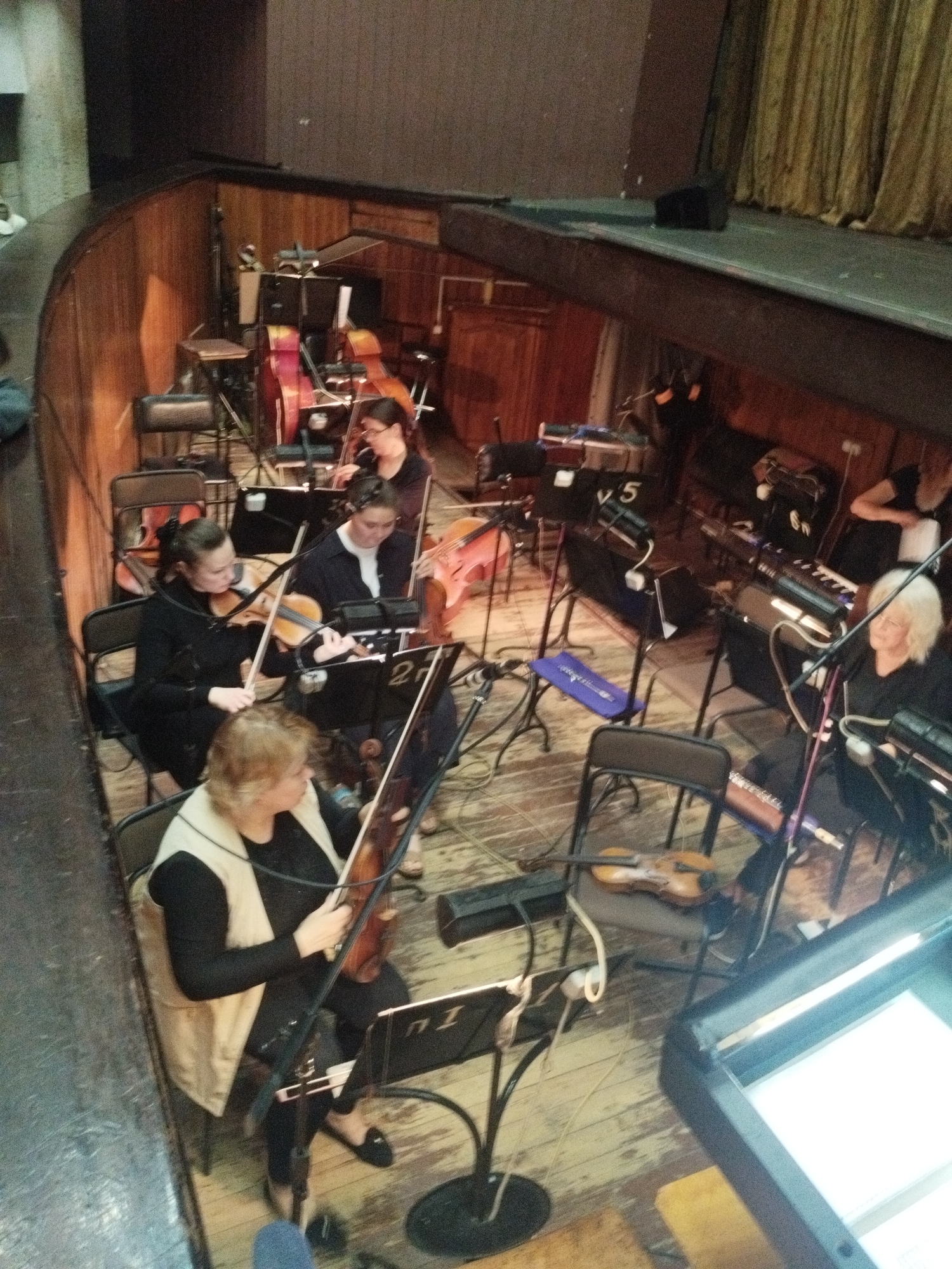 This image depicts an overhead view of an orchestra pit in a theater in Odesa, Ukraine. The pit features dark wood paneling on its curved walls and a light-colored wooden floor. Above the pit, a stage is visible with a dark green or grey floor and gold pleated curtains on the right side.
Nine individuals, predominantly women, are seated within the pit, engaged in playing musical instruments. From bottom left:
* A woman with blonde hair, wearing a black top and a beige vest, is seated and holding a string instrument (likely a viola), facing a music stand marked "П 1".
* Behind her, a woman with dark hair tied back, wearing a black long-sleeved top, is playing a violin, looking at a music stand marked "2".
* Further back and to the left, a woman with dark hair and glasses is playing a cello.
* In the middle-right, a woman with blonde hair is seated at an electronic keyboard, looking at a music stand. Another woman with dark hair is partially visible behind her, also at a keyboard.
* Towards the far right, a woman with dark hair is partially visible playing a larger string instrument, possibly a cello or double bass.
* Other musicians are visible playing string instruments (violins/violas) towards the back and center of the pit.
The pit is equipped with numerous black metal music stands, some bearing numbers such as "2", "5", "6N", and "П 1". Black metal folding chairs are used by the musicians. Multiple microphones on stands, various cables crisscrossing the wooden floor, and sound equipment boxes are present. Several string instruments, including violins, violas, cellos, and a double bass, are visible, along with at least two electronic keyboards. A large red/pink instrument case is positioned in the middle-back. Small lights are affixed to some music stands. A bright screen or monitor is visible in the bottom right corner. A white object or figure is partially visible in the upper left, outside the pit. All musicians appear focused on their instruments and music scores.