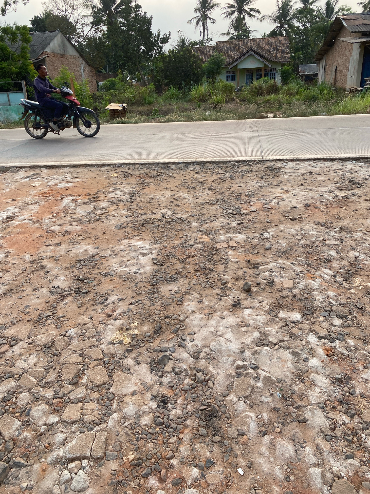 A man rides a black motorcycle on a paved road in Sri Sawahan, Indonesia. The man is wearing a dark long-sleeved shirt and dark pants. He is looking to his left. The road is bordered by a rough, dirt area with many rocks and debris. In the background, there are houses with tiled roofs, palm trees, and other vegetation. The sky is overcast.