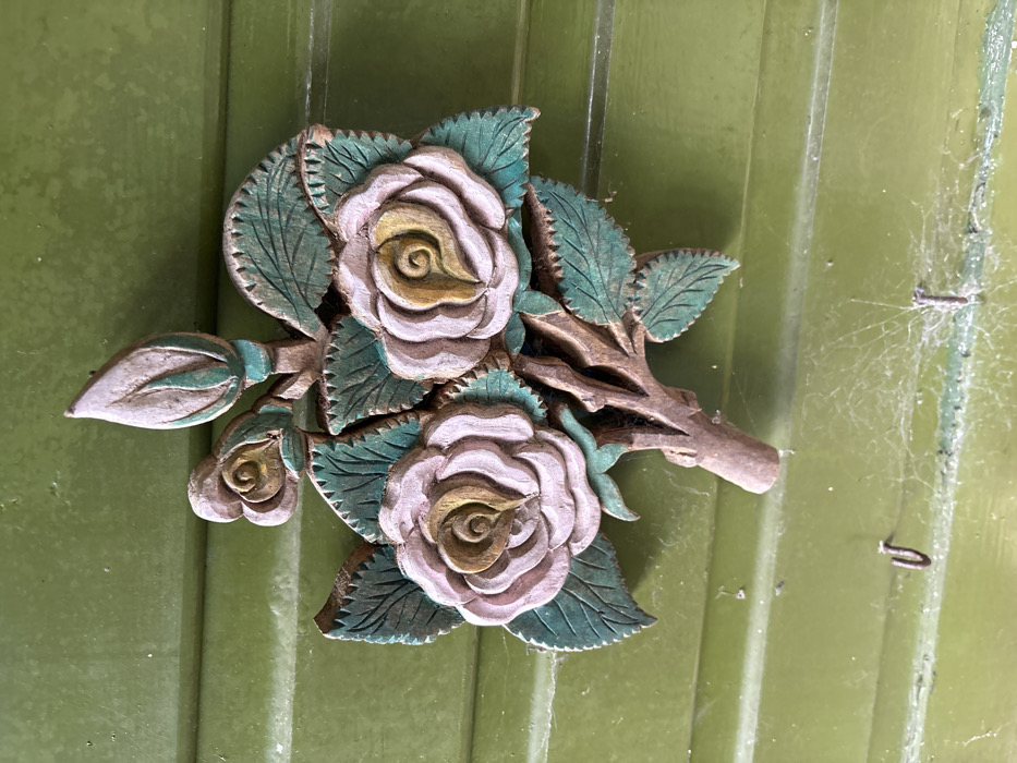 A carved wooden floral decoration, depicting a rose branch with three open roses and one rosebud, is affixed to a vertically paneled, muted green wooden surface. The roses feature pale pink-lilac outer petals with dull yellow-ochre centers. The leaves are painted a dark teal-green with visible veining. The stem is brown, textured to resemble wood. The green wooden background exhibits distinct horizontal panel lines. Cobwebs are present across the lower section of the green surface, obscuring two small, dark, U-shaped hooks located beneath the carving. The scene is situated in Stiperstones, United Kingdom.