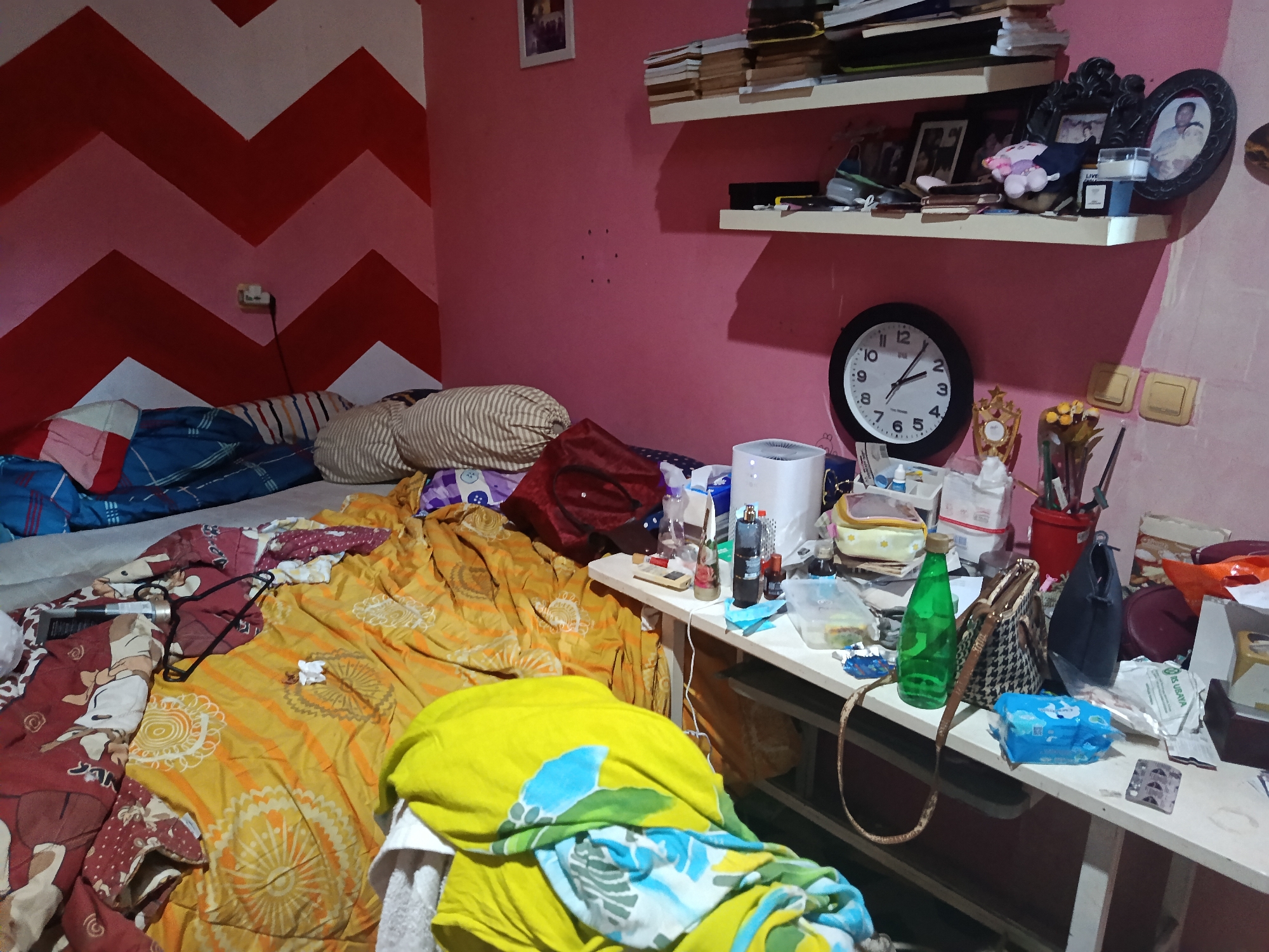 The image depicts a messy bedroom with a bed and a desk. The walls are painted pink, with a red and white chevron pattern on one wall. The bed is covered in a variety of colorful blankets and pillows. On the desk, there are various objects, including a clock, books, a bottle of green liquid, and several bags. The room appears to be cluttered and disorganized. The specific location in the city of Keboananom, Indonesia, is not evident from the image itself.