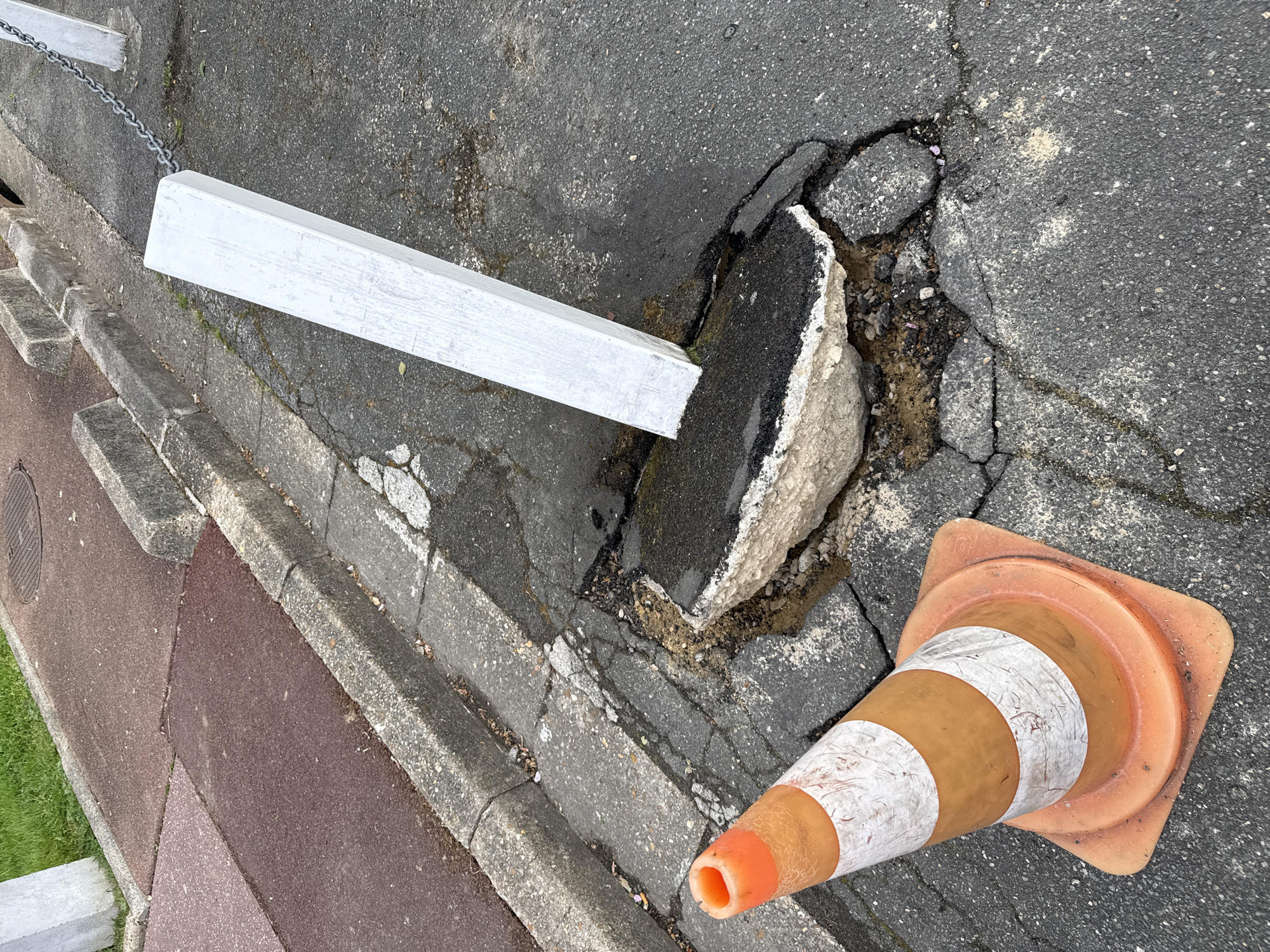 The image shows damage to a road surface in Domont, France. A large chunk of asphalt is broken and dislodged, creating a hazard. A white, rectangular barrier is positioned near the damage, seemingly to cordon off the area. An orange and white traffic cone lies adjacent to the damaged section, likely to alert pedestrians and drivers. The surrounding environment includes a sidewalk with a red paved area, a concrete curb, and a chain railing along the edge. The scene appears to be unattended, with no individuals visible.
