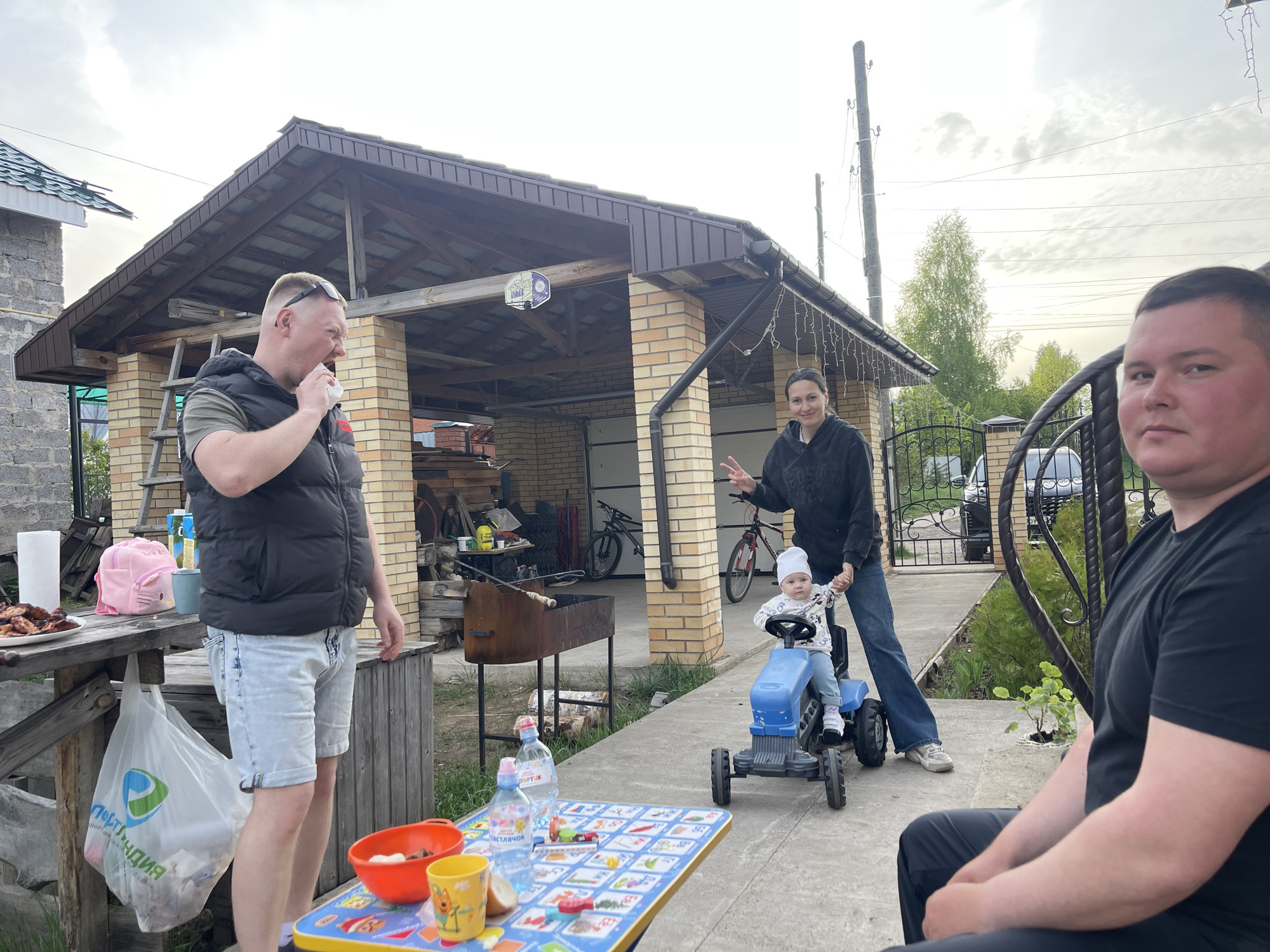 The image depicts an outdoor gathering of people in what appears to be a backyard setting. A man stands on the left wearing a black vest and light blue shorts, holding something to his mouth. A woman stands near the center, holding the hand of a young child who is sitting on a blue toy tractor. Another man is seated on the right side of the frame. In the background, there's a brick building with a garage. There's also a grill, a table with food and drinks, and a fence. The scene is outdoors, under an overcast sky. The location is identified as Chaikovsky, Russia.