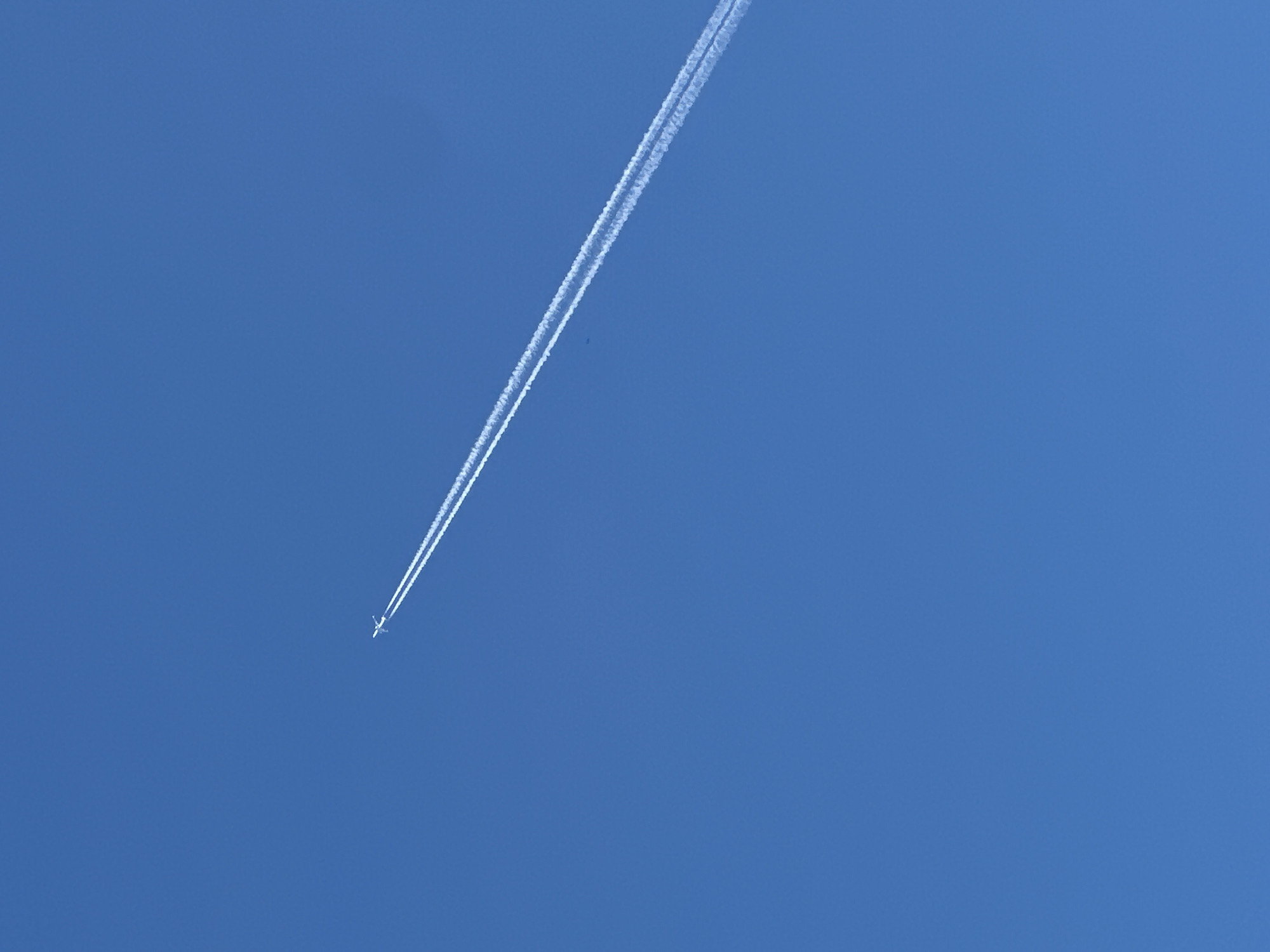 An airplane flies diagonally across a clear blue sky, leaving behind two white contrails. The plane is small in the frame and appears to be moving from the bottom left to the top right of the image. The contrails are distinct and parallel, indicating the path of the aircraft. The sky is a uniform, bright blue, suggesting it is daytime with good visibility. No other objects or elements are visible in the image.