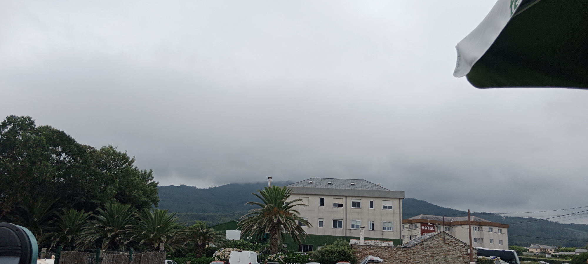 The image depicts a cloudy day in Reinante-praia, Spain. In the foreground, there is a dark green umbrella or canopy, partially obscuring the upper right portion of the frame. To the left, dense green trees and palm trees are visible, with some wooden fencing in front of them.
In the middle ground, there is a multi-story building with a grey roof and a white facade, featuring numerous windows. A palm tree stands in front of this building. To the right of this building, another building is partially visible, with a red sign that reads "HOTEL". In front of the hotel, there is a building constructed with stone.
In the background, a mountain range is covered in dense green vegetation and shrouded in clouds. Electrical wires are strung across the scene. The overall atmosphere is overcast and somewhat muted due to the cloudy weather.
