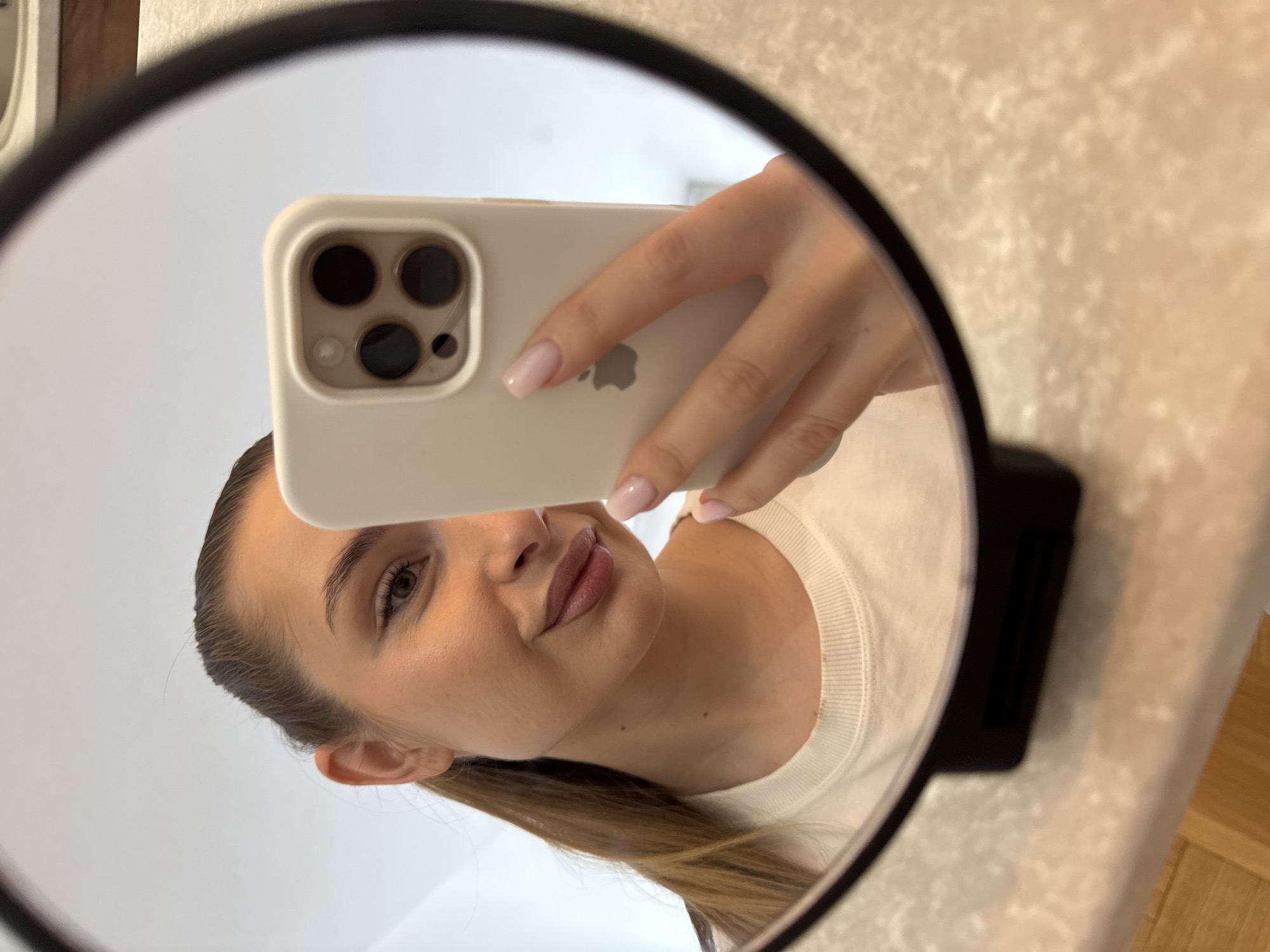 In a mirror selfie, a young woman is reflected, holding a light-colored iPhone with a triple camera system. The smartphone obscures part of her face, with the Apple logo and "iPhone" text visible on its back. The woman has light skin, her brown hair pulled back, and her nails are neatly manicured with a light pink polish. She is wearing a white or off-white ribbed long-sleeved top and displays a subtle, relaxed expression. Her eyes are open, looking into the camera.
The mirror itself is round with a black frame, resting on a stand which is partially visible at the bottom right. The background of the reflection includes a plain, light-colored wall or ceiling behind the woman. To the right of the mirror, a textured, light-toned surface, possibly a countertop or wall, is visible, suggesting an indoor setting. A hint of a wooden floor can be seen at the bottom right of the frame. The lighting appears bright and even, suggesting daytime. The overall scene captures a casual moment in Budva, Montenegro.