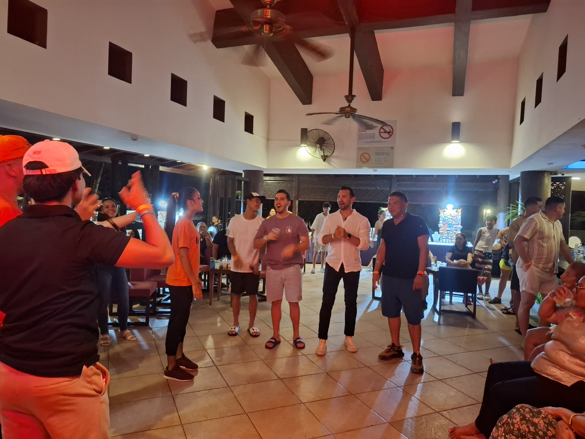 The scene is a well-lit indoor space, likely a resort lobby or common area in Playa El Agua, Venezuela. A group of about 10 people are gathered centrally, seemingly engaged in an activity led by a person wearing a white cap, black shirt, and salmon-colored shorts with his back to the camera, directing the others with animated gestures. Several other people are scattered around the room, some seated and watching, others standing. The building's interior features a high ceiling with dark wood beams and ceiling fans. Signage regarding no smoking is visible on the wall. The floor is tiled, and the overall atmosphere suggests a casual social gathering.