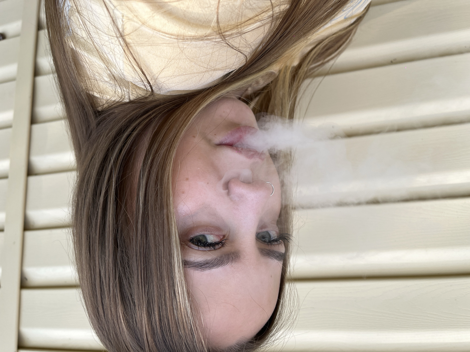 A young woman with light brown hair and green eyes is exhaling a cloud of vapor. She is wearing a nose ring. The background consists of light beige horizontal siding. The location is Novopokrovskaya, Russia.