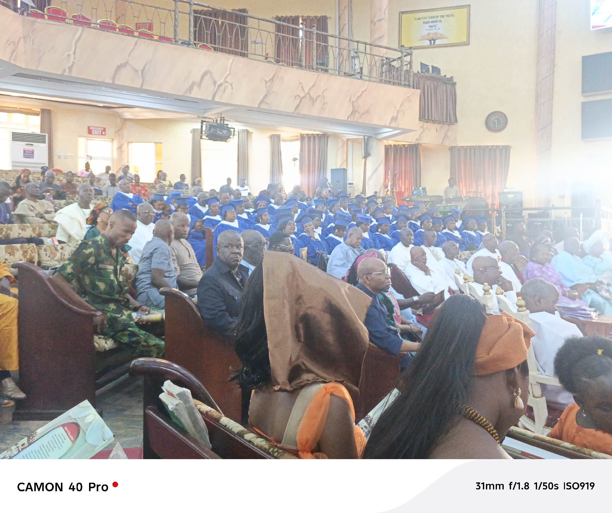 The image shows a large group of people seated in what appears to be a church or auditorium. Many are wearing blue graduation gowns and caps, suggesting a graduation or recognition ceremony. The crowd consists of people of varying ages, with some wearing traditional attire. The seating arrangement is in rows of pews. A man in military camouflage is visible in the left foreground. There are framed pictures on the wall and text above the crowd, including "SANCTIFY THEM BY THE TRUTH" and "YOUR WORD IS TRUTH." The building's interior has beige walls and a balcony overlooking the seated crowd. The location is in the city of Unknown, Unknown.