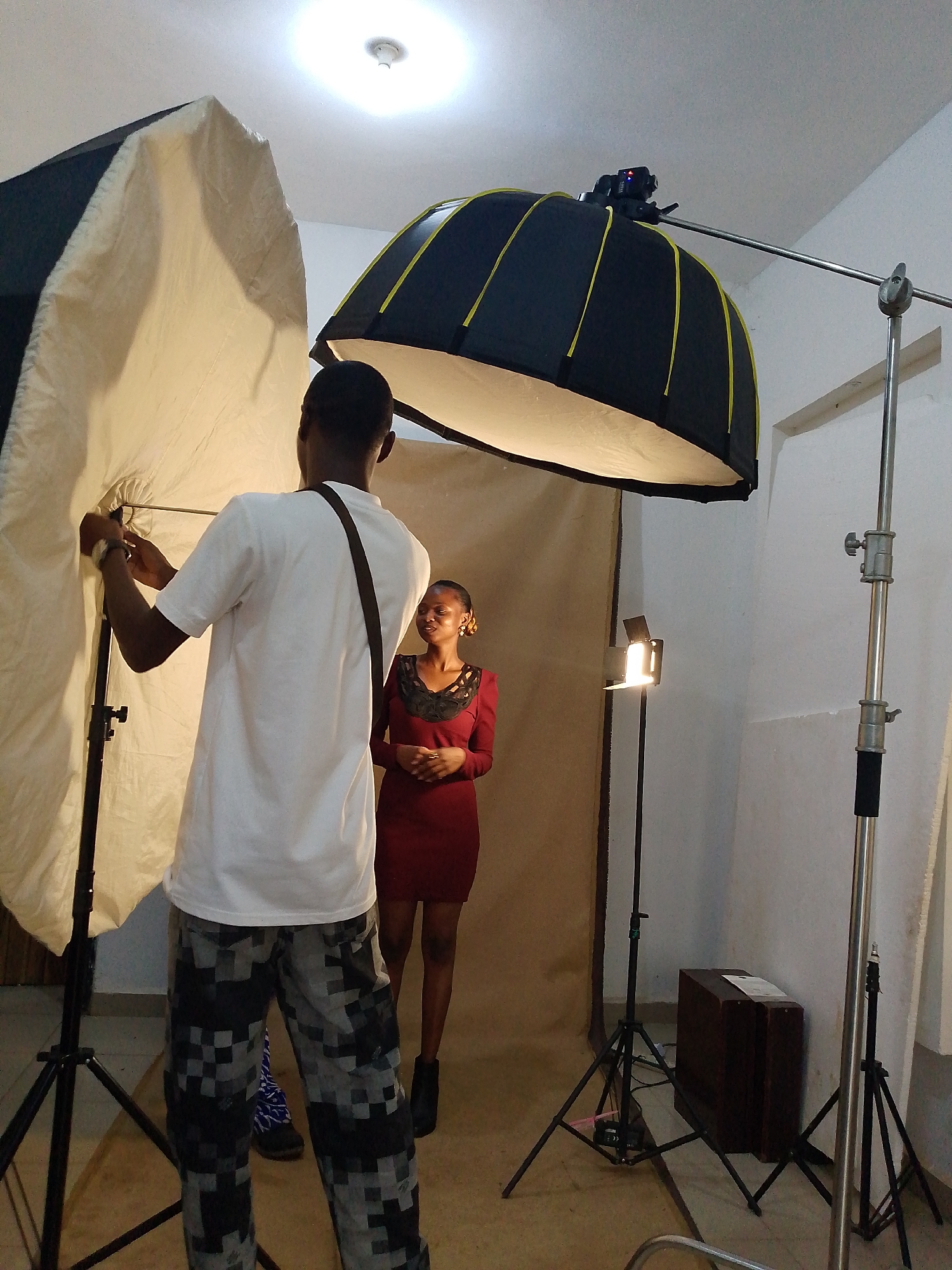 In a room at an Unknown location in Unknown, a man adjusts a large white umbrella light stand while standing beside a woman in a red dress. She has her hands clasped together and is looking at the man. A smaller lamp is visible behind her on a black stand. Behind them is a brown backdrop. To the right is another, overhead umbrella light on a stand, and a dark brown chest is visible to the right. The room has white walls and a light-colored floor. The man wears a white shirt and patterned pants.