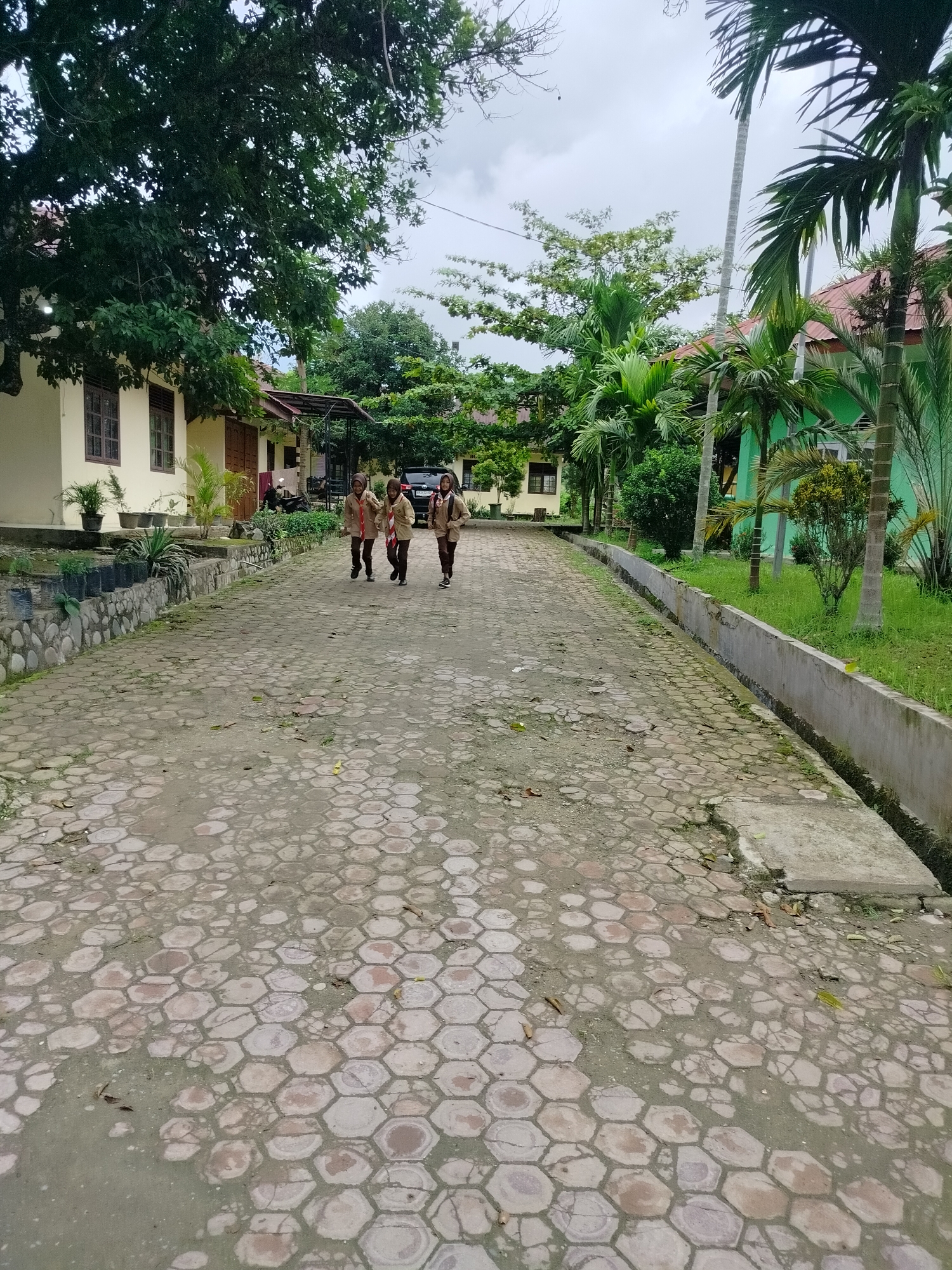 Three young people, likely students, dressed in tan uniforms with red scarves, are walking down a paved path. They are positioned in the middle ground of the image, moving away from the viewer. The path is constructed of hexagonal concrete pavers, some of which appear worn or damaged. To the left of the path are buildings with light-colored walls and brown window frames, interspersed with potted plants and greenery. To the right, there is a concrete drainage channel running alongside the path, bordered by grass and lush vegetation, including palm trees and other tropical plants. In the background, more buildings and trees are visible under an overcast sky. The scene appears to be an outdoor area of a school or educational institution in Purwodadi, Indonesia, given the architectural style, vegetation, and the uniforms worn by the individuals.