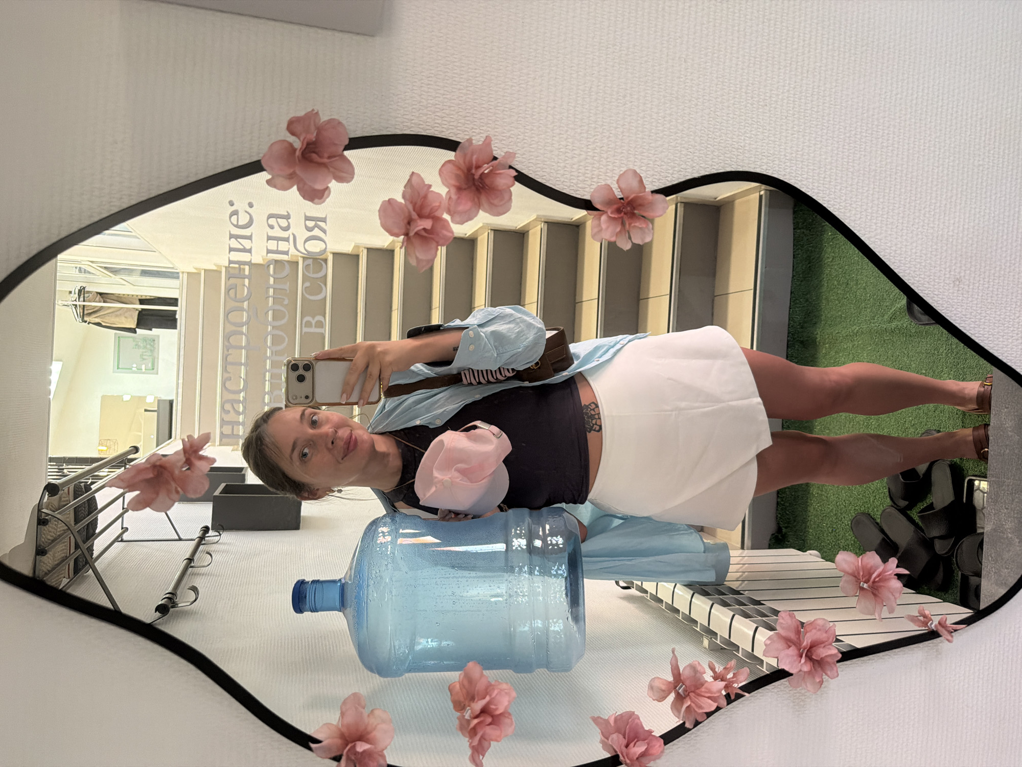 The image is a mirror selfie, presented sideways, capturing a young woman taking her picture indoors. The subject, a woman with light brown hair, is seen from the chest down, holding a smartphone to capture her reflection. She wears a light blue button-down shirt unbuttoned over a dark top, a white mini-skirt, and sandals. A brown shoulder bag is slung across her body, and a pink baseball cap rests on her midriff. A tattoo is visible on her left side. She has a slight smile and is looking towards the phone.
The mirror has a dark, possibly black, rounded frame, and is decorated with several pink artificial flowers attached to its surface or rim. In the lower left foreground, a large, clear blue water bottle with a darker blue cap lies horizontally on a white textured surface, which also reflects in the mirror.
The background, reflected in the mirror, reveals an indoor setting. Visible elements include a flight of stairs, a section of green artificial turf, a white and black striped platform or bench, and a clothing or shoe rack holding dark slippers. Further in the reflection, what appears to be a hallway or another room is visible with a light fixture and a framed picture on the wall. The lighting suggests daytime.
Prominently displayed on the mirror's surface in white Cyrillic text is the phrase: "настроение: положена в себя," which translates to "Mood: invested in myself."