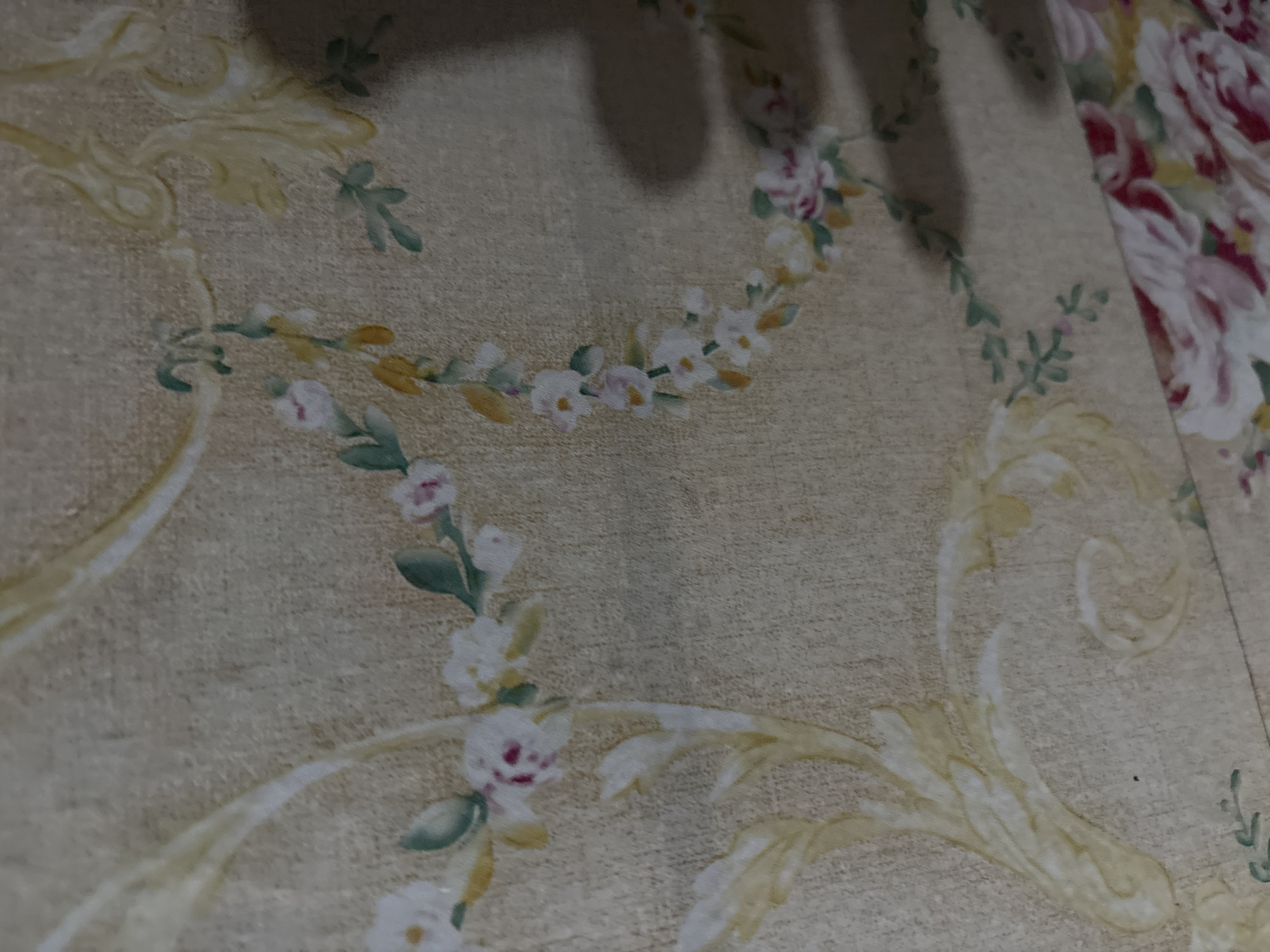 The image displays a close-up, angled view of floral wallpaper with a textured, beige background. The wallpaper features an intricate pattern of light yellow, swirling botanical motifs and delicate clusters of white and pink flowers with green leaves. Some of the flowers are partially obscured or blurred, suggesting the image is not perfectly in focus. In the lower left portion of the frame, a hint of more vibrant, large pink and white flowers suggests a larger floral arrangement on the same wallpaper or a contiguous fabric. The lighting creates subtle shadows on the textured surface, adding depth to the pattern. The specific location in Danawinangun, Indonesia, is not discernible from the visual information presented in the image.