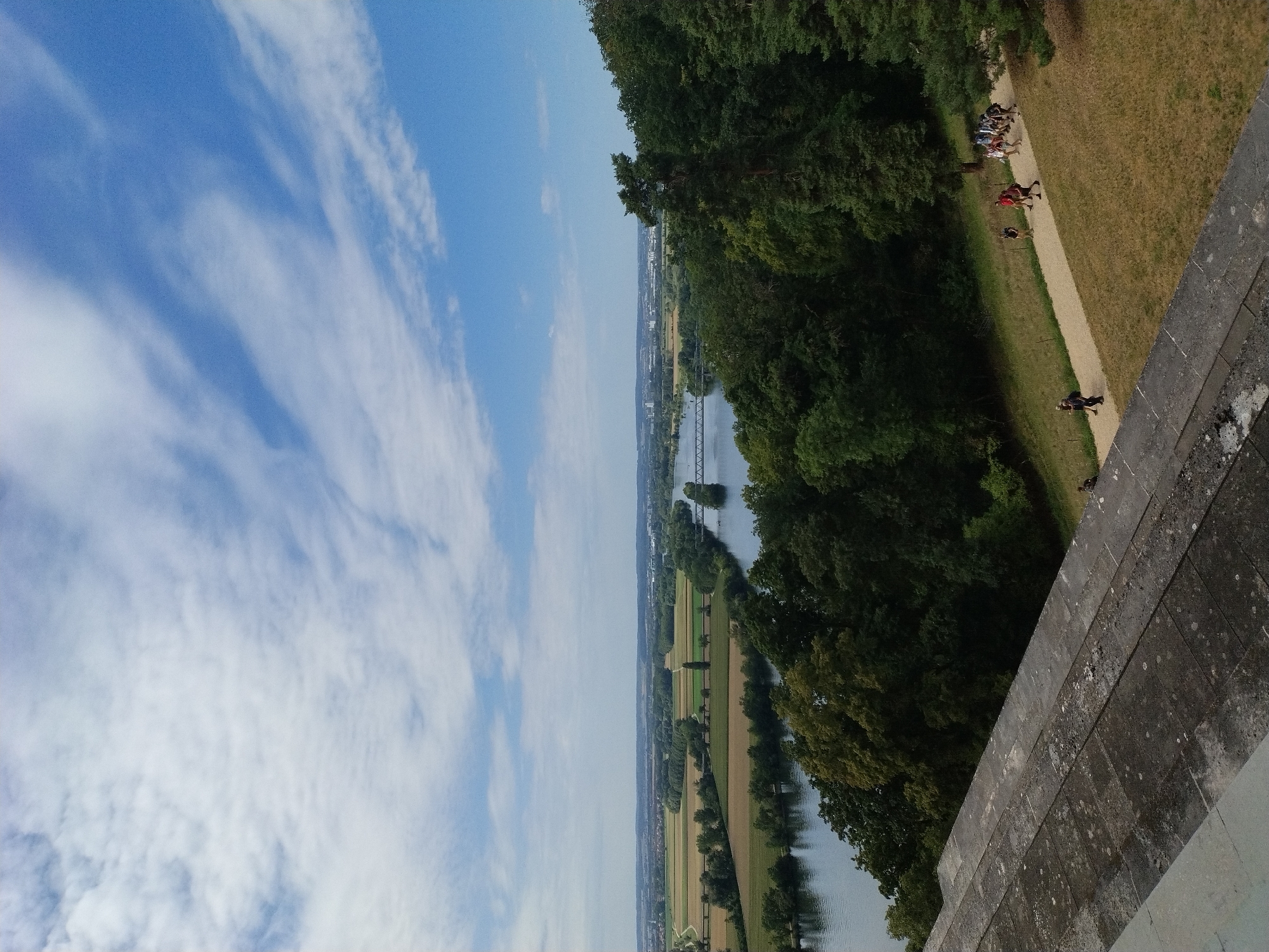 A wide-angle, slightly downward-facing shot captures a panoramic view of the Danube River and surrounding landscape in Donaustauf, Germany. In the foreground, a stone platform with visible grout lines and moss patches occupies the bottom left corner. Beyond the platform, a pathway meanders through a grassy slope dotted with trees. A group of approximately ten people, dressed in casual attire, are walking along the pathway, some in small clusters, others individually. The pathway leads towards a dense forest of deciduous and coniferous trees that borders the river. The Danube River, wide and calm, flows through the middle ground, reflecting the sky. On the opposite bank, agricultural fields in shades of green and golden brown are visible, with a road or path running alongside the river. In the far distance, a city skyline with buildings and a bridge crossing the river can be seen. The sky is a vibrant blue with scattered white, wispy clouds. The overall scene depicts a sunny day in a scenic natural and semi-urban environment.