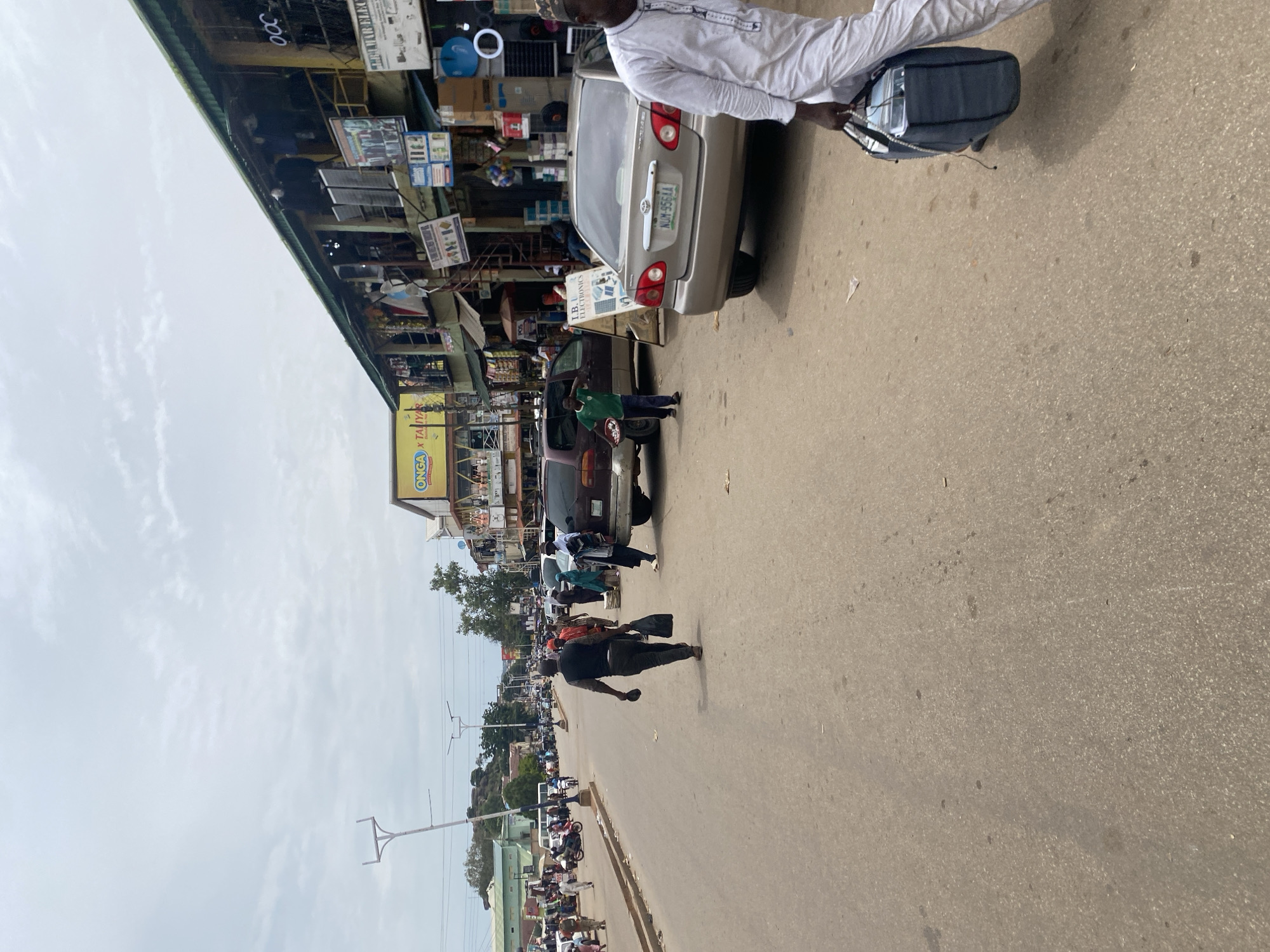 The image captures a busy street scene in Jos, Nigeria, likely a marketplace. In the foreground, a man in a white robe walks away from the camera, carrying a dark blue bag. To his left, a beige sedan with the license plate NUM 956 AA is parked. Further down the street, several people are walking and interacting. On the right side of the street, a two-story building with shops is visible, featuring various signs and goods displayed outside. On the left side of the street, there is a line of vehicles and people. The sky is overcast. The overall impression is one of an active urban environment with commercial activity.