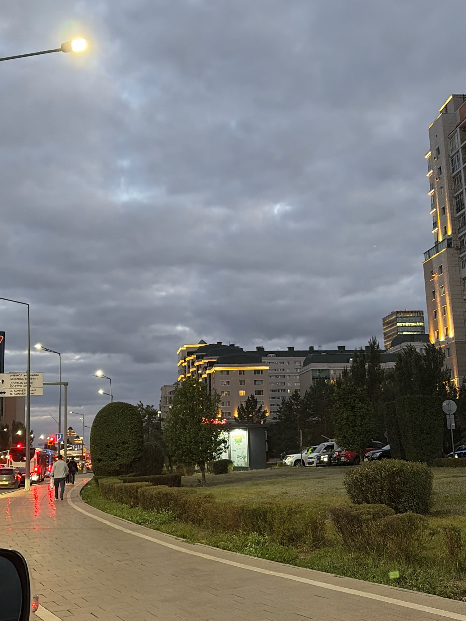 The image depicts a street scene in Astana, Kazakhstan, on a cloudy day. A lamppost illuminates the overcast sky on the left side of the frame. In the background, several multi-story buildings are visible, some featuring illuminated architectural details. On the right, a tall, modern building is prominently lit with yellow lights.
In the foreground, a man walks away from the viewer on a paved walkway. To the left of the walkway, a street is lined with cars and a bus, with red brake lights reflecting on the wet pavement. Trees and manicured bushes form a green median between the walkway and the street. A information kiosk stands near the edge of the green space. Several cars are parked in the background, near the buildings. The overall atmosphere is one of a moderately busy urban environment during twilight or overcast weather.