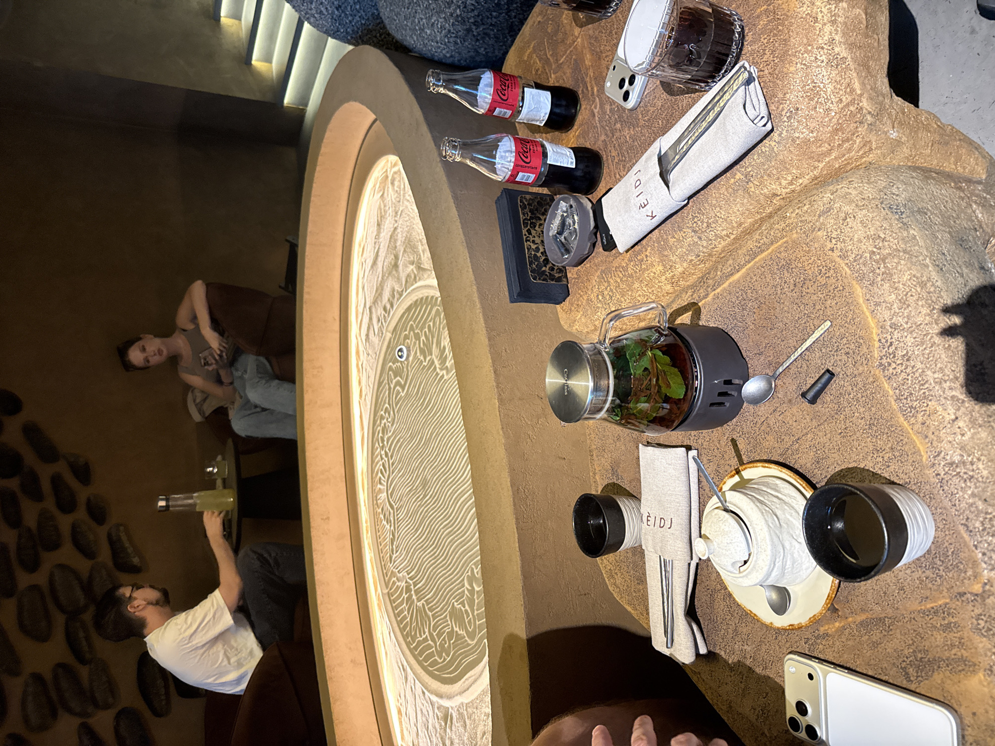 In the foreground, a rough, textured stone table holds various items. A white ceramic sugar bowl with a lid sits on a saucer, accompanied by a small spoon. Next to it is a black and white striped ceramic cup filled with a dark liquid. A metal teapot with a glass carafe filled with a beverage and green leaves is also present, along with a set of chopsticks resting on a linen napkin with the word "KEIDJ" printed on it. Two glass bottles of Coca-Cola are on the table, as is a dark napkin holder with a speckled design. A mobile phone with a white case is visible on the left side of the table. In the mid-ground, a large, circular sand garden with intricate wave-like patterns is illuminated from above. Two people are seated in the background, with a man in a white t-shirt and jeans on the left, holding a glass bottle. A woman in a gray top and blue jeans is seated on the right, looking towards the camera. The environment appears to be an indoor seating area with dim lighting and a textured wall adorned with dark, oval-shaped decorative elements. The overall impression is that of a relaxed dining or lounge setting.