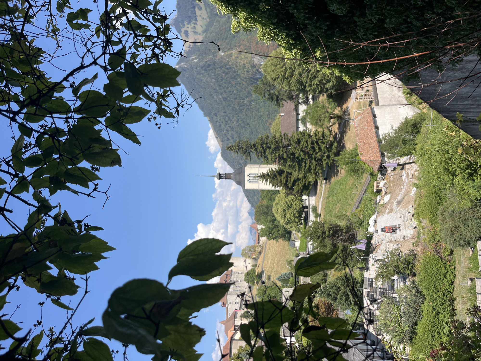 An outdoor, high-angle, eye-level shot captures a quaint European town nestled at the foot of a mountain. In the foreground, lush green foliage frames the scene, creating a natural border. Beyond the greenery, a pale stone church with a dark steeple stands prominently, its spire reaching towards the bright blue sky dotted with white clouds. To the left of the church, a historical castle complex, with its terracotta roofs and stone walls, sits perched on a hillside, suggesting a long and rich history. The landscape is a harmonious blend of architecture and nature, with rolling hills, dense forests, and the imposing mountain forming a picturesque backdrop. A construction site is visible in the middle ground, with heavy machinery and piles of rubble indicating ongoing work. The overall impression is one of serene beauty, with the juxtaposition of ancient structures and modern development.