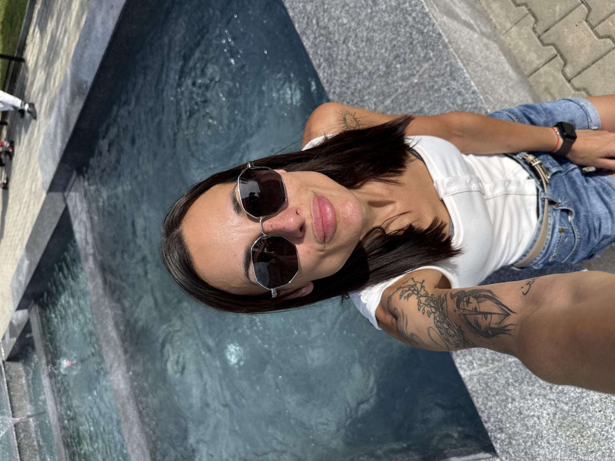 A woman is taking a selfie by a pool. She is wearing sunglasses, a white tank top, and denim shorts. She has tattoos on her arm. The pool is made of dark stone with blue water. In the background, two people are walking on a paved area. The scene appears to be outdoors on a sunny day.