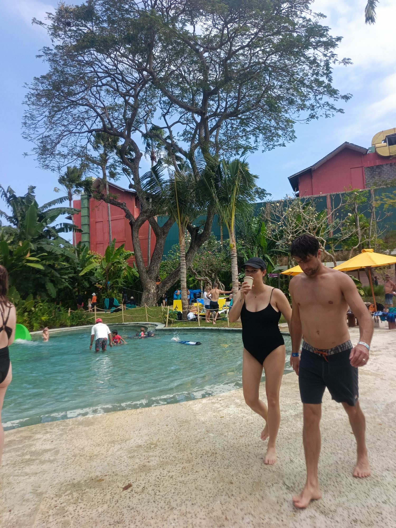 The image depicts a sunny day at a water park in Kuta, Indonesia. In the foreground, a couple is walking on a sandy path beside a swimming pool. The woman, wearing a black swimsuit and a black cap, is holding a cup to her mouth. The man, shirtless and wearing blue patterned swim trunks, walks beside her. In the pool, several people are swimming and wading, including a person in a red shirt. Lush green tropical vegetation and trees surround the pool area, with a large, red building in the background. Yellow umbrellas provide shade in the distance. The sky is clear and blue.