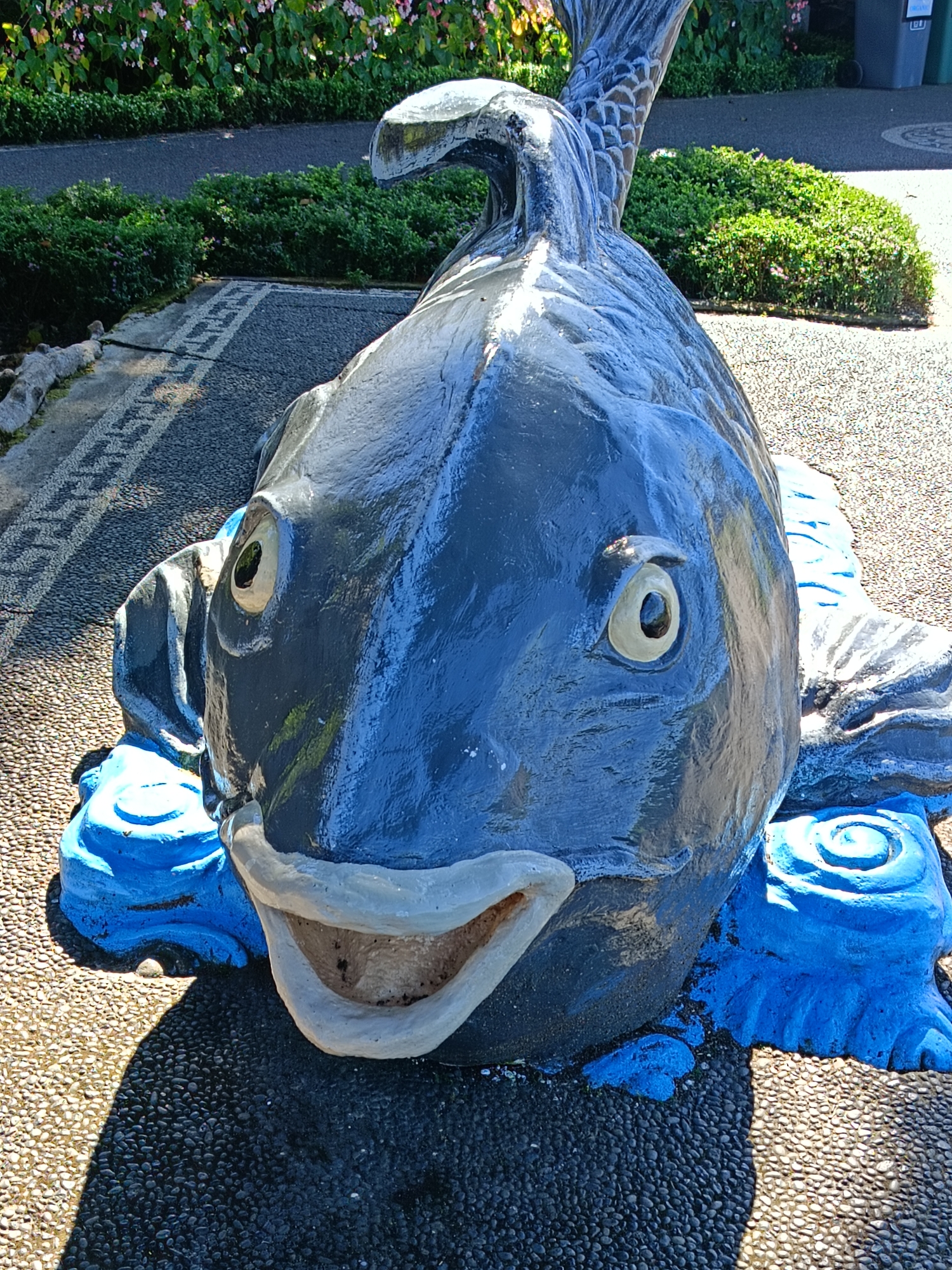 The image shows a close-up, eye-level view of a large, dark blue, fiberglass fish sculpture. The sculpture is situated outdoors on a pathway composed of dark and light-colored pebbles. The fish has a wide, open mouth with a pale gray interior and a light gray outline. Its eyes are large and round with a yellowish-beige iris and a dark pupil. The sculpture is supported by blue wave-like structures at its base. In the background, there are neatly trimmed green bushes and a colorful flower bed with pink and white blossoms. Further in the background, part of a gray trash bin is visible. The scene appears to be a public space, possibly a park or garden area, within the city of Baturiti, Indonesia. The lighting suggests it is daytime, with distinct shadows cast by the sculpture.