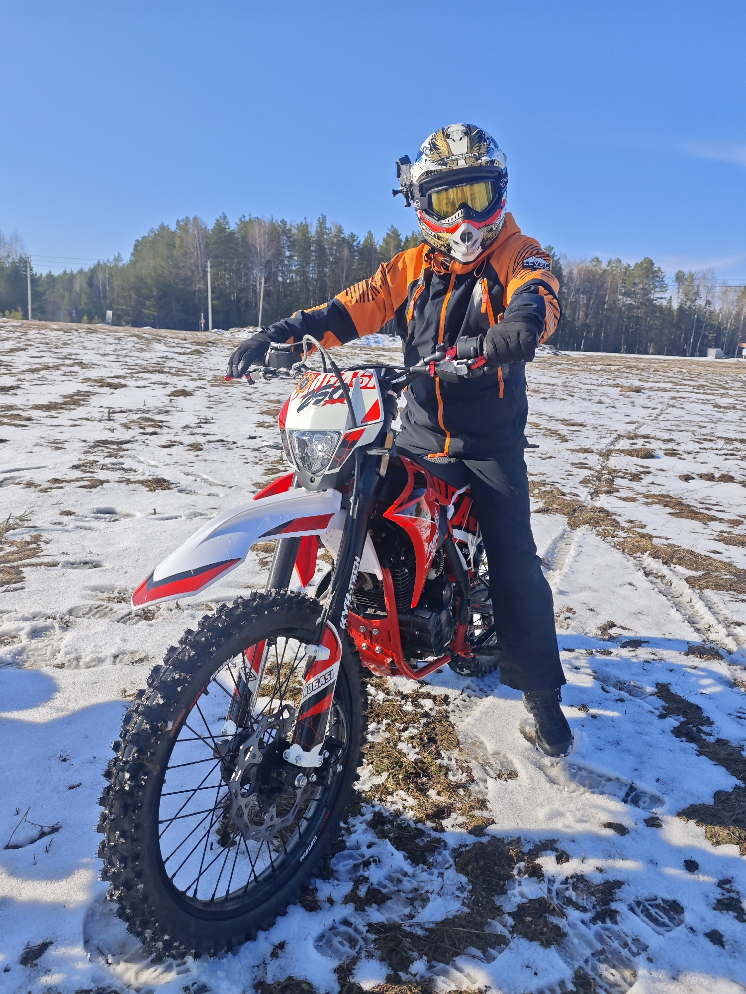The image shows a person on a red and white dirt bike in a snowy field. The person is wearing a helmet with goggles, an orange and black jacket, and black pants. The field is covered with patches of snow and dry grass. In the background, there is a line of trees. The photo was taken in Прикамский, Russia.