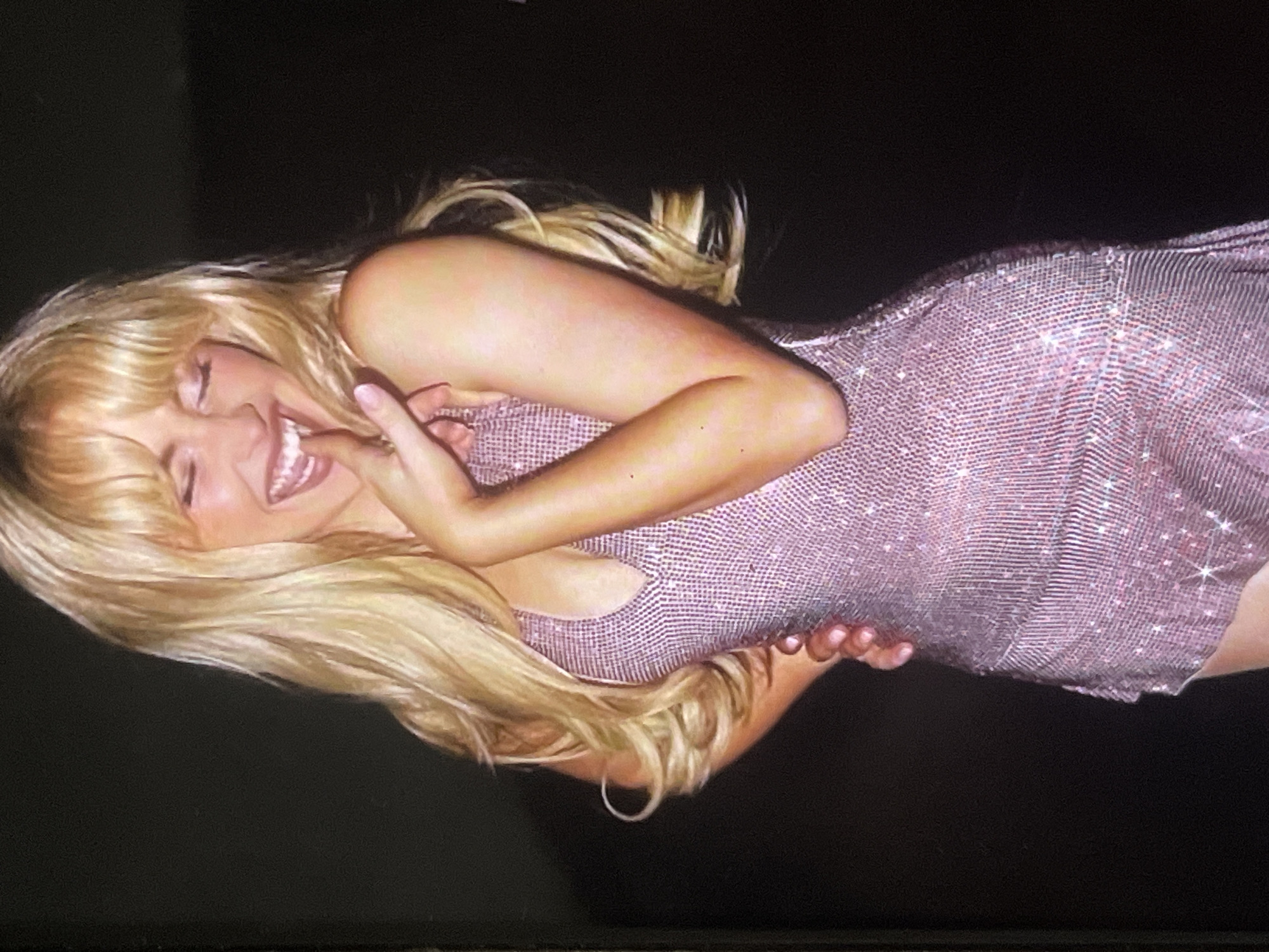 The image, oriented sideways (rotated approximately 90 degrees counter-clockwise), features a young woman with long, voluminous blonde hair and bangs, caught in a moment of joyful laughter. Her eyes are closed or squinted due to her broad smile, and her right hand is brought up towards her mouth, partially covering it. She is wearing a shimmering, short, sleeveless dress in a light pink or lavender hue. The dress is made of a reflective fabric, possibly mesh or sequins, which catches light and creates a sparkling effect. It is body-hugging and appears to be above knee-length.
The background is uniformly dark, suggesting a low-light environment such as an evening event or an indoor setting. Artificial light illuminates the woman and her dress, accentuating the sparkle and her features. Her blonde hair flows around her, with some strands visibly curled. The dominant emotion conveyed is happiness and amusement. No text is visible in the image. This scene is situated in Poltava, Ukraine.