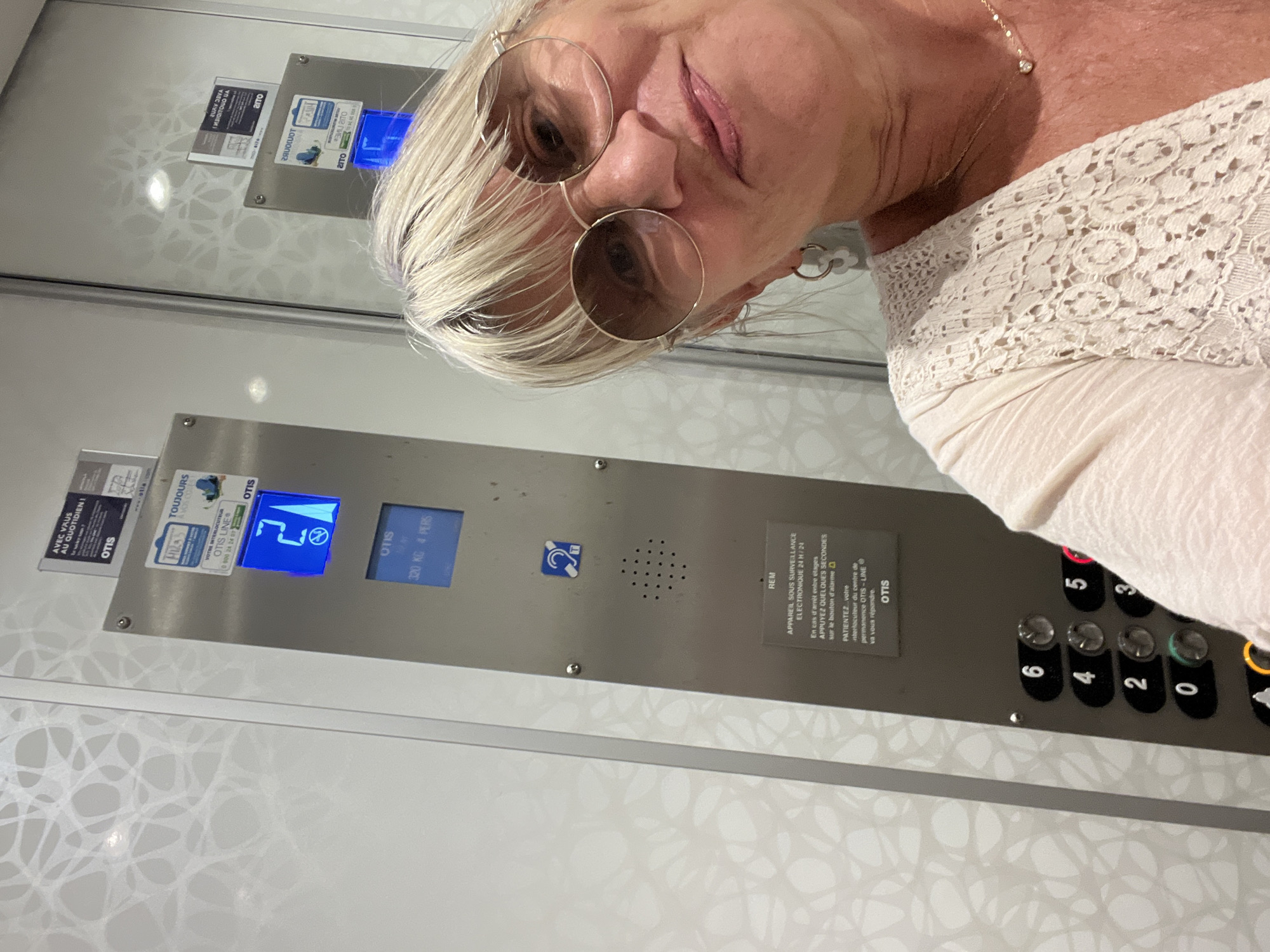 A woman with blonde hair and sunglasses is taking a selfie in an elevator. She is wearing a cream-colored lace top and a delicate necklace. The elevator panel is visible, showing buttons numbered 0 through 6, as well as a digital display indicating the number 2. There are also various stickers and labels on the elevator panel, including "OTIS". The elevator walls are white with a subtle patterned design. The scene appears to be in Palavas-les-Flots, France, likely at a hotel or residential building given the elevator.