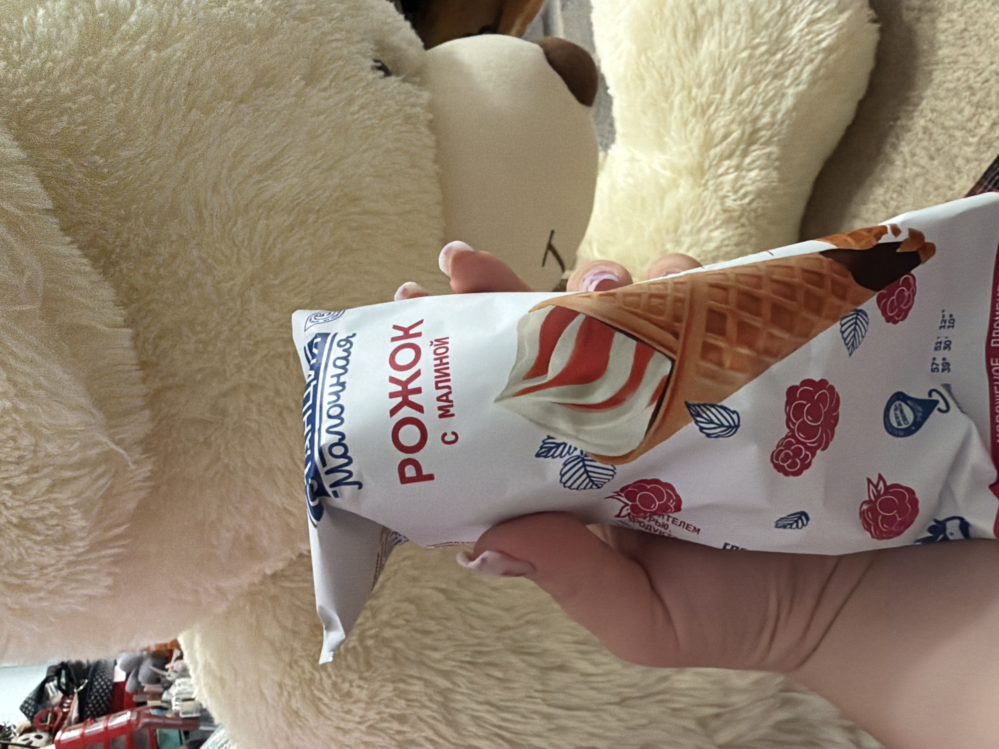 In this close-up shot, a hand is holding a wrapper for an ice cream cone, featuring a waffle cone with swirling red and white ice cream, drizzled with chocolate. The wrapper is primarily white with red and blue illustrations of raspberries and leaves. Some text is visible on the wrapper, including "Молочная", "РОЖОК", and "С МАЛИНОЙ" in Cyrillic script, indicating it is a raspberry ice cream cone. The wrapper also displays nutritional information and other text in smaller font. The background is dominated by the fuzzy texture of a large, light-colored stuffed animal, possibly a bear, adding a soft, cozy feel to the scene. The lighting suggests it is daytime indoors. The presence of the ice cream and the soft toy evokes a sense of comfort and relaxation. The overall impression is of a casual moment, possibly at home, enjoying a sweet treat.