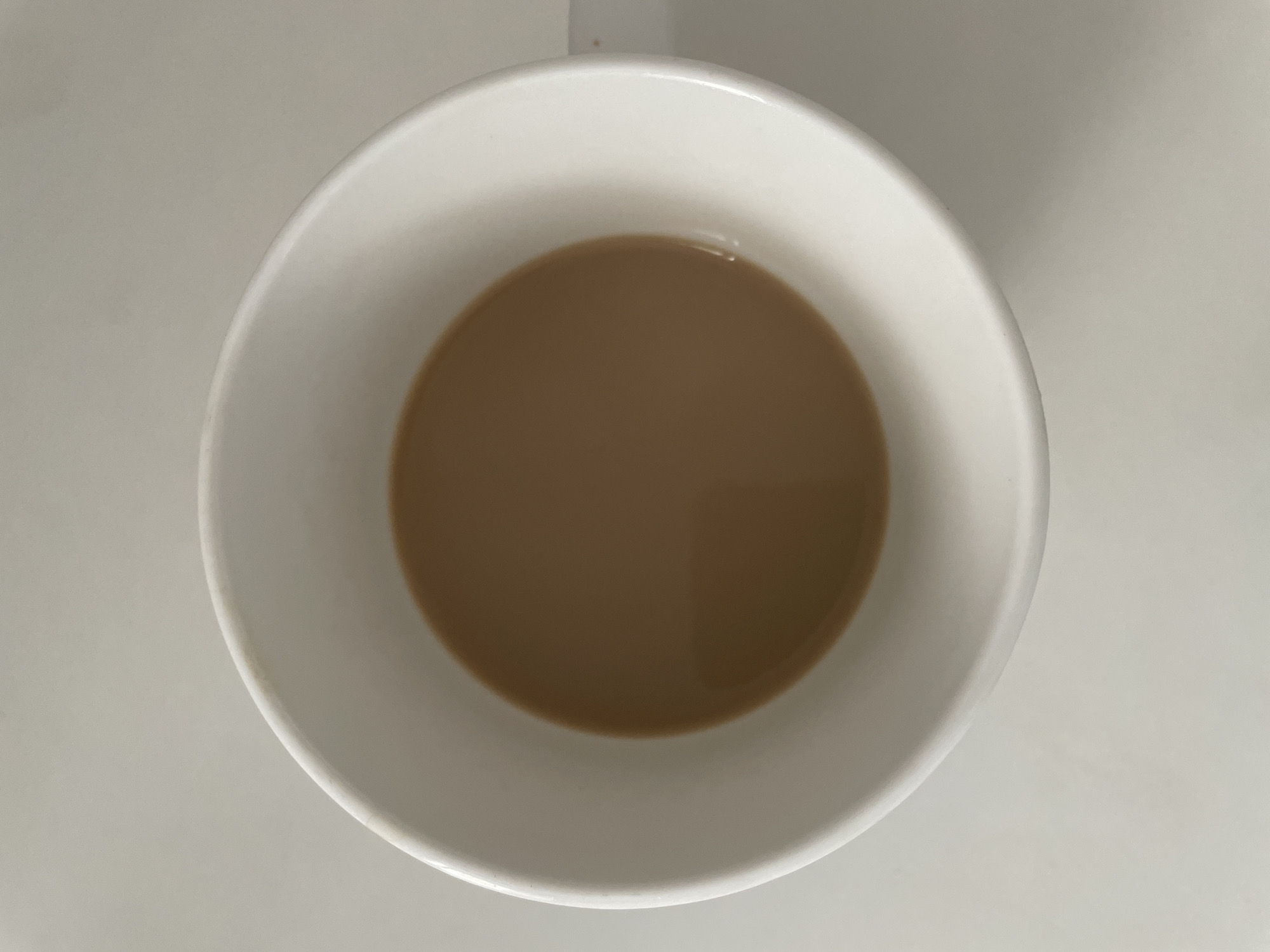 The image presents a top-down view of a white ceramic mug, centrally framed against a plain, light grey or off-white surface. The mug contains a light brown liquid, consistent in color with coffee or tea mixed with milk, filling approximately one-quarter of its volume. The surface of the liquid is smooth and still. The interior and exterior of the mug are uniformly white, though subtle, light brown discoloration or staining is visible along portions of the upper rim, particularly on the top-left edge. The surrounding surface is unadorned and lacks any identifying features. No individuals, additional objects, actions, or contextual elements are depicted. There are no visual cues or landmarks within the image that allow for the determination of a specific geographical location, including the city of Емельяново, Russia.