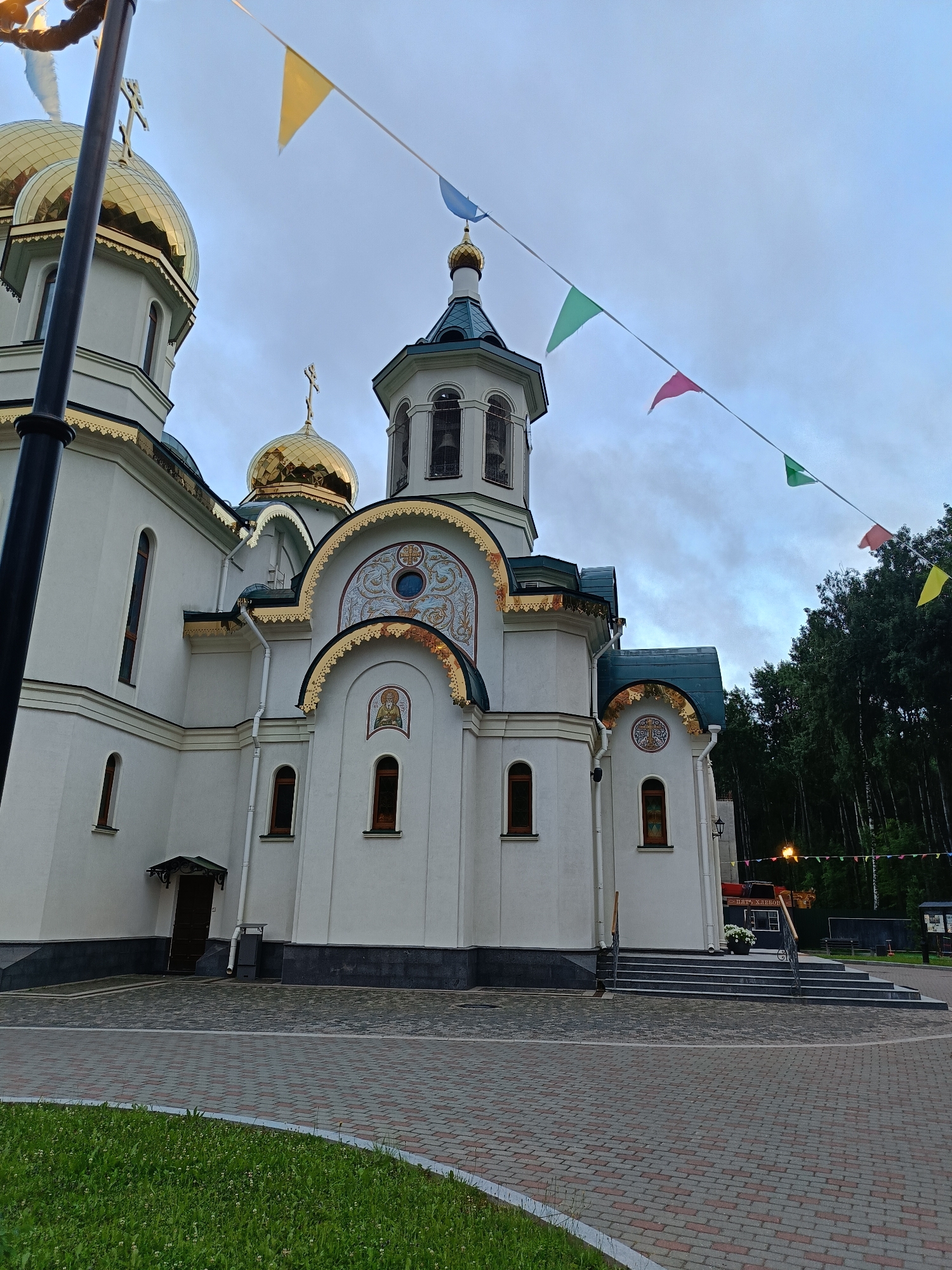This is a ground-level, wide-angle, slightly upward-tilted shot of a white Orthodox church in Saint Petersburg, Russia. The church features multiple golden domes and a teal roof. A string of multi-colored triangular flags stretches across the upper portion of the frame, against a cloudy, grey sky. The foreground consists of a patterned brick walkway and a patch of green grass. The church's entrance is visible on the left, and a set of stairs leads to an entrance on the right side. The background is dominated by a dense line of trees. There are no people visible in the image. The lighting suggests it is either early morning or late evening.