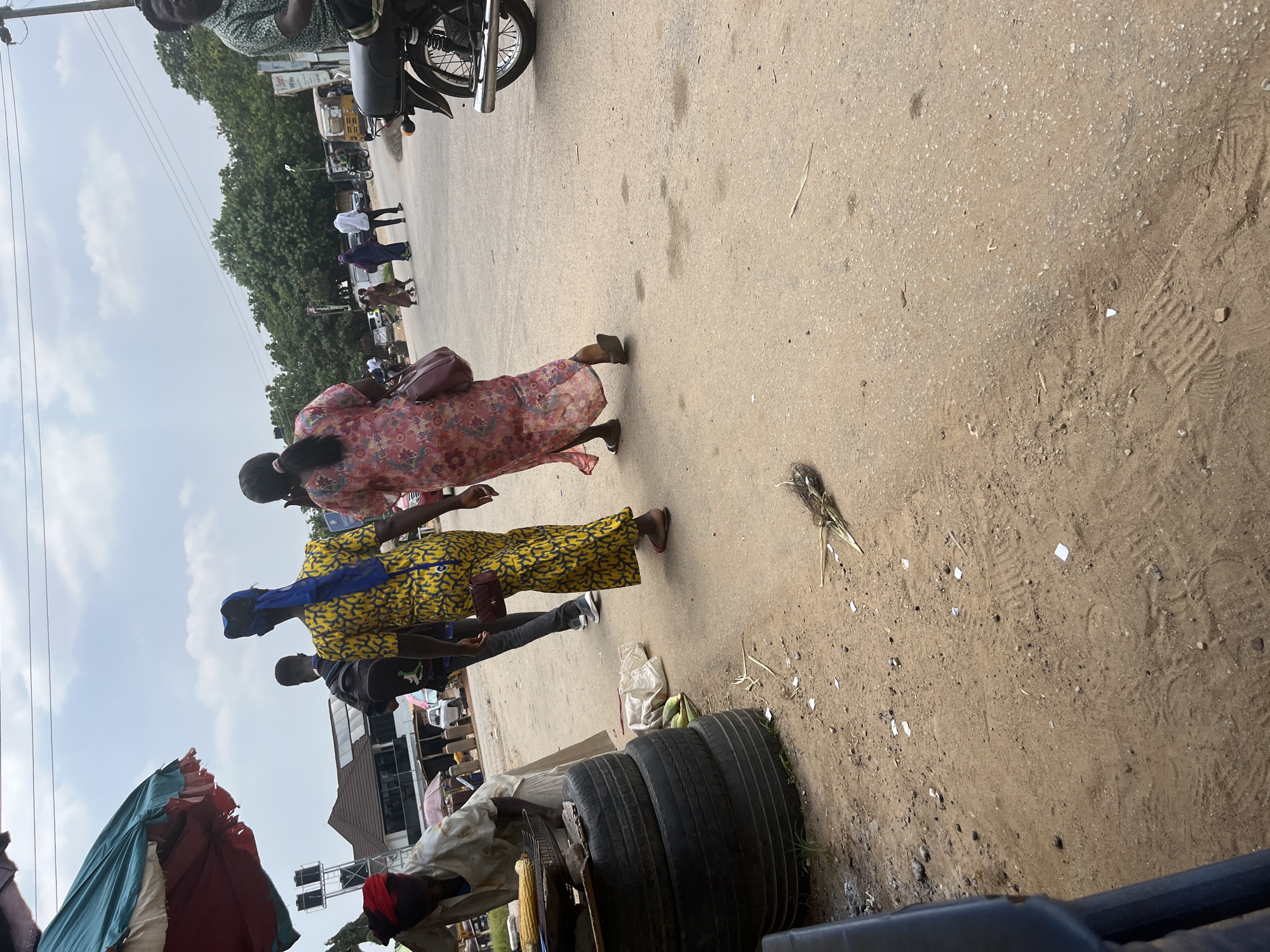 The image captures a street scene in Abuja, Nigeria, likely during the daytime given the bright lighting. The perspective is from a slightly elevated angle, looking down at the ground and a group of people walking on a sandy, paved road.
In the foreground, a woman in a vibrant, floral-patterned dress walks from left to right, carrying a burgundy handbag over her shoulder. Behind her, another person in a yellow and blue patterned dress is visible, also walking in the same direction. A third individual, wearing dark clothing and a blue head covering, is walking just behind the person in yellow. The ground is a mix of dirt and sand with scattered debris, including what appear to be dried plant material and small pieces of paper.
Further in the background, more people are walking, and several vehicles, including a motorcycle and what looks like a commercial van, are parked or in motion. Trees line the side of the road, suggesting an urban or semi-urban environment. A market stall with colorful tarps is partially visible on the left side of the image. Stacked tires are also present on the right side of the road, near the foreground.
The overall atmosphere seems to be that of a busy public space, possibly a market area, with people going about their daily activities. The weather appears to be clear, with some clouds in the sky. No specific text is clearly legible in the image.