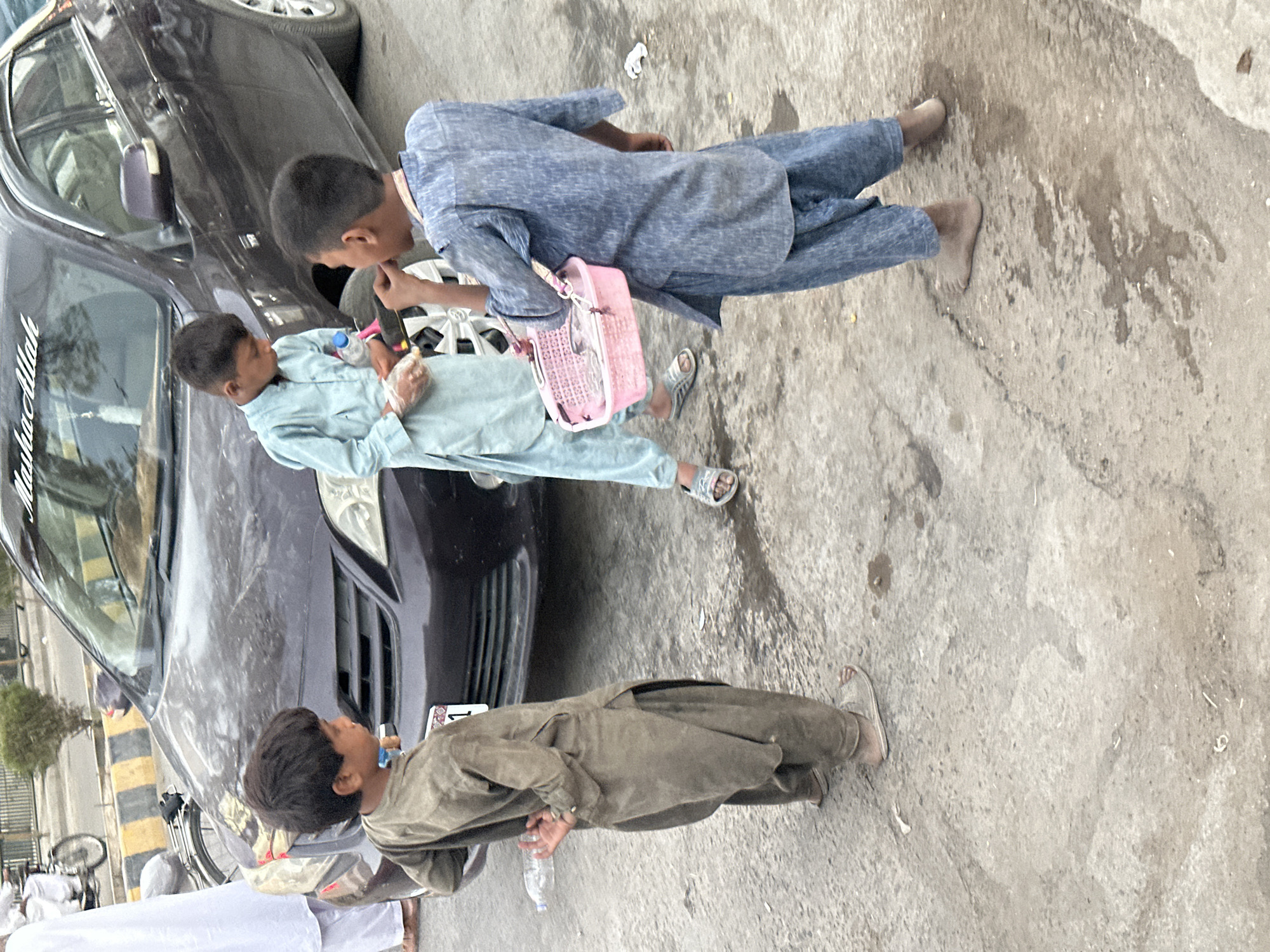 In Quetta City Tehsil, Pakistan, three young boys are observed near a dark-colored sedan. The boy on the left, wearing a faded olive-green shalwar kameez, stands with his back to the viewer, looking towards the right. He has dark hair and is barefoot. To his right, another boy in a light blue shalwar kameez is facing the sedan. He has a clear plastic bottle in his left hand and a small white package in his right. He is also barefoot. The third boy, on the far right, is wearing a blue shalwar kameez and is seen from the back, facing the other two boys. He is holding a pink plastic basket with a strap over his shoulder, and appears to be eating something. His feet are bare. The boys are standing on a rough, light-colored concrete surface with some dark stains. A portion of the sedan's front grille and a license plate are visible. In the background, a street with a curb and some vegetation can be seen.