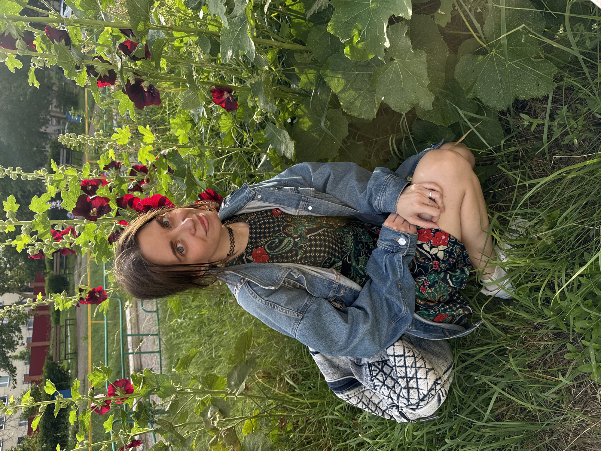 A young woman with brown hair and striking blue eyes is crouched in the grass, surrounded by tall green plants with large leaves and dark red flowers. She is wearing a dark, patterned dress, a denim jacket, and white sneakers. A patterned bag rests beside her. Her left hand is resting on her knee, and she's wearing a ring. The woman is looking directly at the camera with a relaxed expression. The scene appears to be outdoors, likely in a garden or park, with hints of urban buildings and playground equipment in the background, suggesting it could be a residential area. The lighting suggests it is daytime, likely afternoon, with a warm, natural light illuminating the scene. The overall mood is peaceful and serene. There is no visible text in the image.
