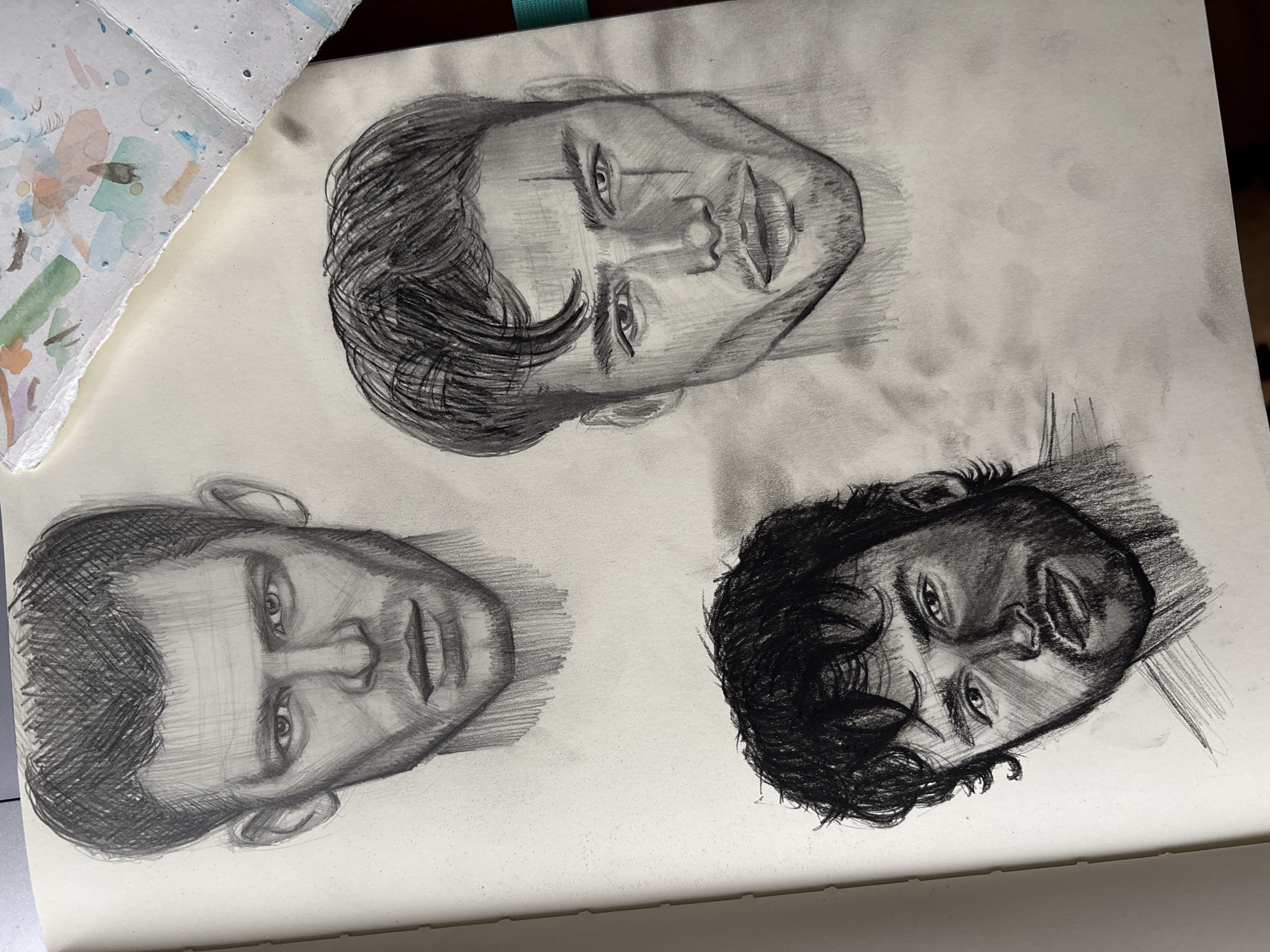 The image shows a sketchbook page with three pencil portraits of male faces. The top face has short, dark hair styled to one side, a prominent brow, and a defined mouth. The face in the bottom left has a shorter haircut, a broader face, and softer features. The face in the bottom right has dark, curly hair and a more angled face. The background consists of a white sketchbook page. In the top left corner, there is a partially visible watercolor palette with various colors mixed together. There is no evidence of Izhevsk, Russia, or any location information in the image.