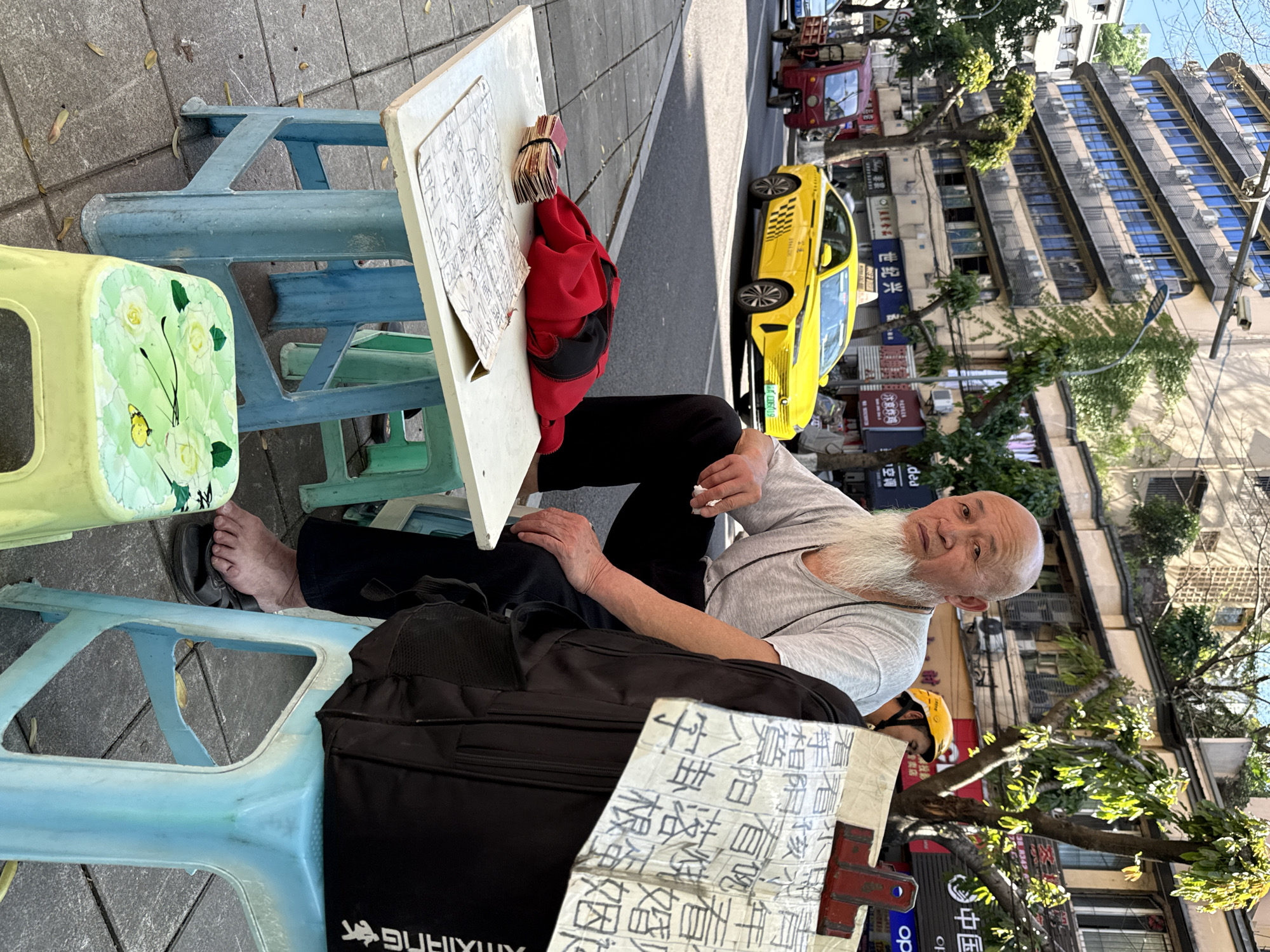 Here is a forensic description of the image:
**Scene:**
The image shows a man sitting on a small blue plastic stool on a sidewalk. He is seated at a small white table with documents and money on it. The scene appears to be in an urban area, potentially in the city of Beibei, China, considering the Chinese script visible in the background and on the table documents.
**People:**
The main subject is an elderly man with a long white beard. He is wearing a grey t-shirt and black pants. He has one foot out of his sandal. Another person wearing a yellow hard hat is near the table.
**Objects:**
* A small white table with documents written in Chinese.
* A stack of money.
* A red bag on the table.
* A black bag next to the stool.
* Two plastic stools (one blue, one green).
* A yellow taxi on the street.
* A red tricycle.
* Buildings and trees in the background.
**Actions:**
The elderly man is sitting down, holding what appears to be a small piece of paper or a cigarette. He is looking toward the camera.
**Environment:**
The setting is an urban street. There is a sidewalk where the man is seated. Buildings and shops are visible in the background, with signs written in Chinese. The street appears to be active with traffic. The time of day appears to be daytime, under natural light.