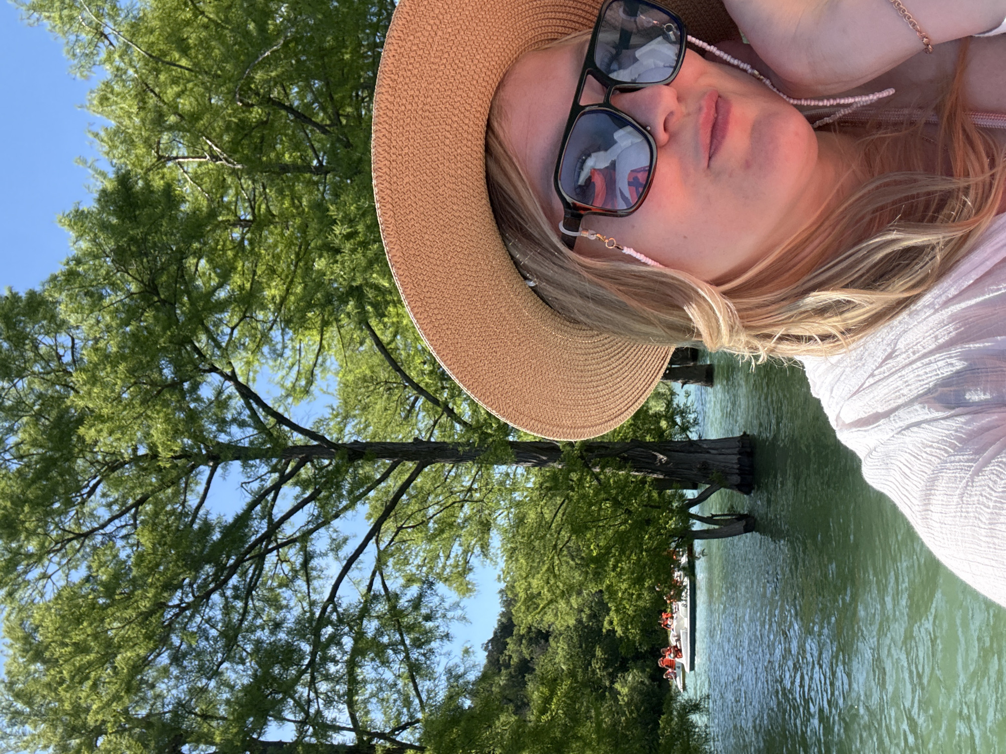 The image captures a sunny outdoor scene in Sukko, Russia, centered around a person relaxing by a body of water. The setting features lush green trees and clear blue skies, suggesting pleasant weather.
In the foreground, a person, likely female, is partially visible from the chest up, positioned as if lying down or reclining. They have blonde hair, fair skin, and are wearing black-framed sunglasses and a light-colored, possibly white, textured top. A wide-brimmed, light brown straw hat partially covers their head. A delicate beaded chain is attached to their sunglasses, and a light-colored bracelet is on their wrist. Their lips are slightly parted, showing a soft pink hue.
The background reveals a serene body of water, possibly a lake or river, extending into the distance. Several large, dark tree trunks are partially submerged in the water. Further back on the water, multiple small white boats are visible, each carrying passengers who appear to be wearing orange life vests. The surrounding landscape is rich with verdant foliage, dominated by tall trees with numerous branches and leaves.
The activity depicted is one of leisure and enjoyment, with the person relaxing outdoors near the water, while other people are seen boating. The bright sunlight and clear skies indicate it is daytime, likely a pleasant morning or afternoon. The overall atmosphere conveys tranquility and recreation. There is no visible text in the image.
