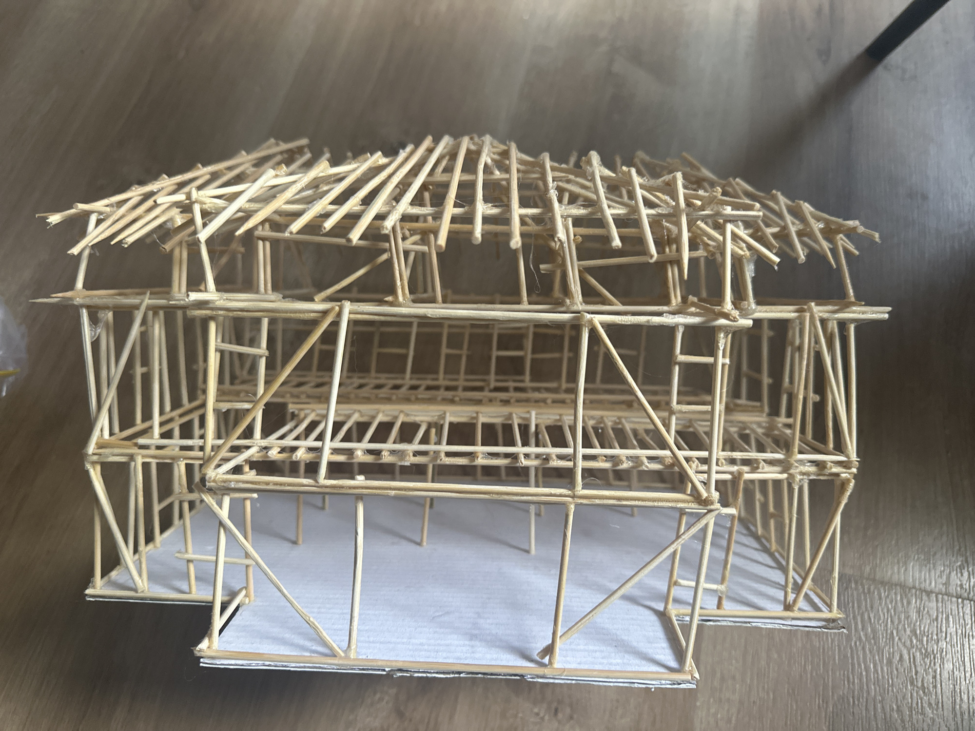 The image shows a two-story architectural model constructed from wooden sticks, possibly toothpicks or skewers, and glue. The model sits on a white surface, likely paper or cardstock, which rests on a light brown wooden floor. The model depicts a building with a rectangular base, two levels of open framework for walls, and a loosely constructed roof made of the same wooden sticks laid across the top. There are no people visible in the image. The model appears to be handcrafted and may be a school project or architectural prototype. There is no evidence to suggest the image was taken in Sofia, Bulgaria.