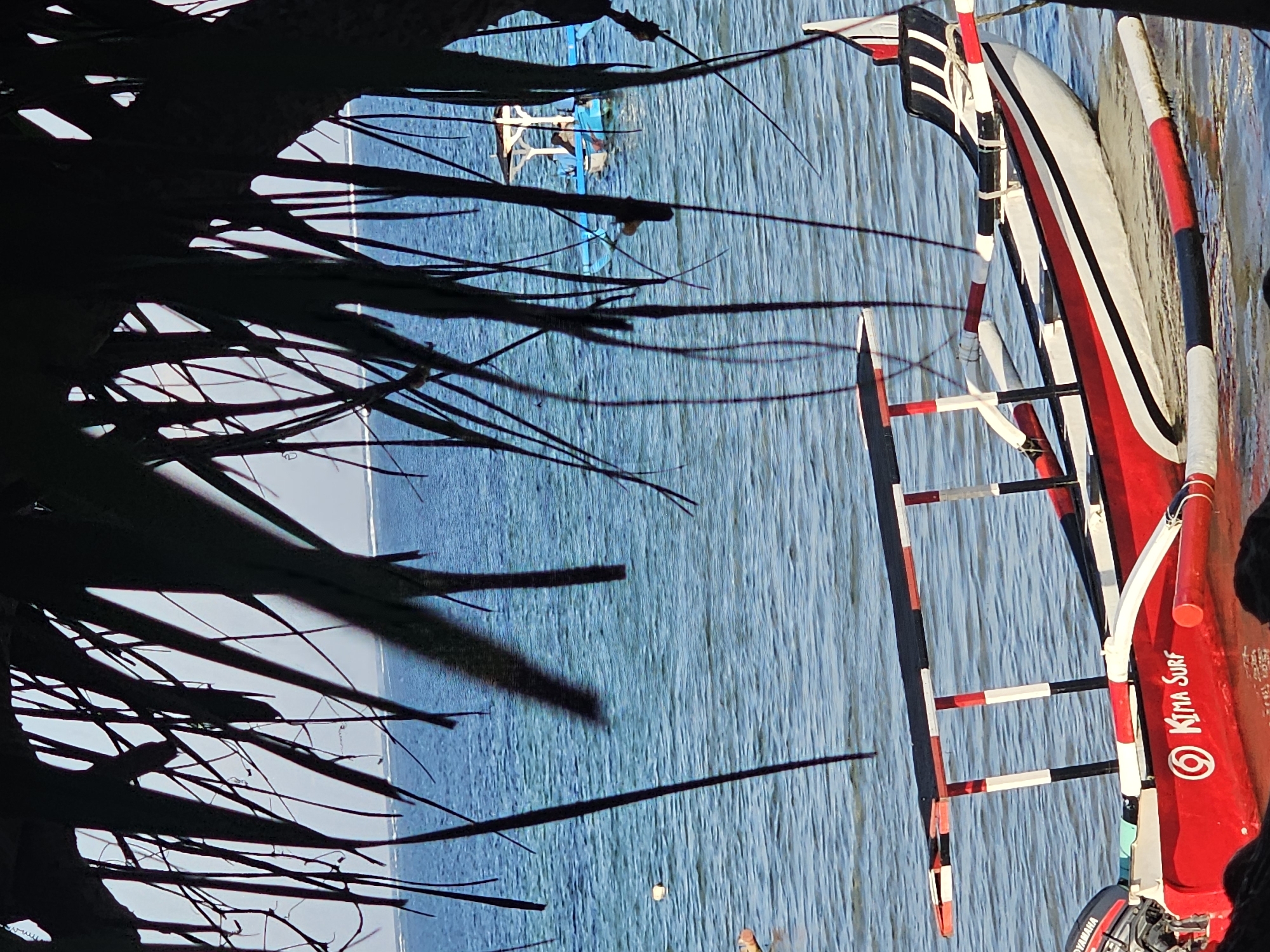 The image depicts a serene ocean scene, likely in a coastal area of Denpasar, Indonesia. The foreground is dominated by a vibrant red and white outrigger canoe, adorned with black and white accents on its canopy supports and seating. The canoe is partially submerged in calm, blue water, with a Yamaha outboard motor attached to its stern. The name "Kima Surf" is prominently displayed on the side of the canoe, along with a circular logo.
In the background, another, smaller boat with a white canopy and blue hull is visible further out on the water. The ocean stretches to the horizon, meeting a hazy sky. The scene is framed by the dark, silhouetted fronds of palm leaves, suggesting the vantage point is from under a thatched roof or a cluster of palm trees, common in beachside locations. The water appears shallow near the shore, with the outrigger canoe resting close to what seems to be the seabed. The overall impression is one of tranquility and a typical day at a tropical beach.