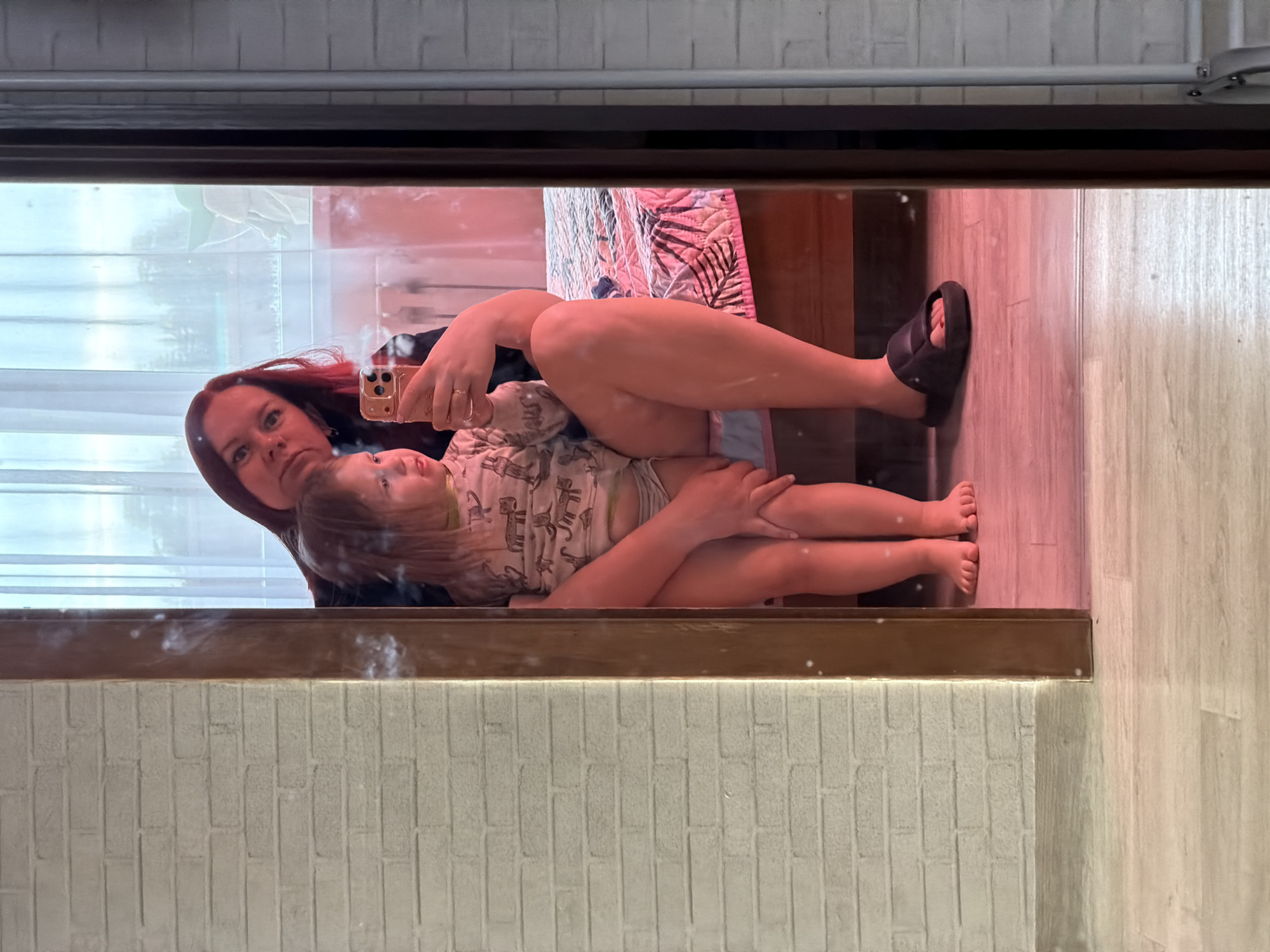An indoor mirror selfie captures an adult female with reddish-brown hair and a young child. The female is holding a gold-colored smartphone in her left hand, featuring a ring on her left ring finger, while her right arm is positioned around the child. She is wearing a dark top and a single black slide sandal on her right foot. The child, with blonde hair, is wearing a light-colored top with animal patterns and light bottoms, looking towards the left side of the frame. Both individuals are reflected sitting on or adjacent to a bed covered with a pink and dark leaf-patterned quilt. The immediate environment reflected includes a light-colored laminate floor, and to the left of the dark brown mirror frame, a light-colored faux-brick textured wall is visible. In the background, through a window with sheer white curtains, vegetation and a partial view of a light green, possibly curved or conical, architectural element are discernible. A radiator is also partially visible below the window. The reflected scene exhibits a reddish ambient light cast. The specific location within the city of Chaikovsky, Russia, is not discernible from the provided image.