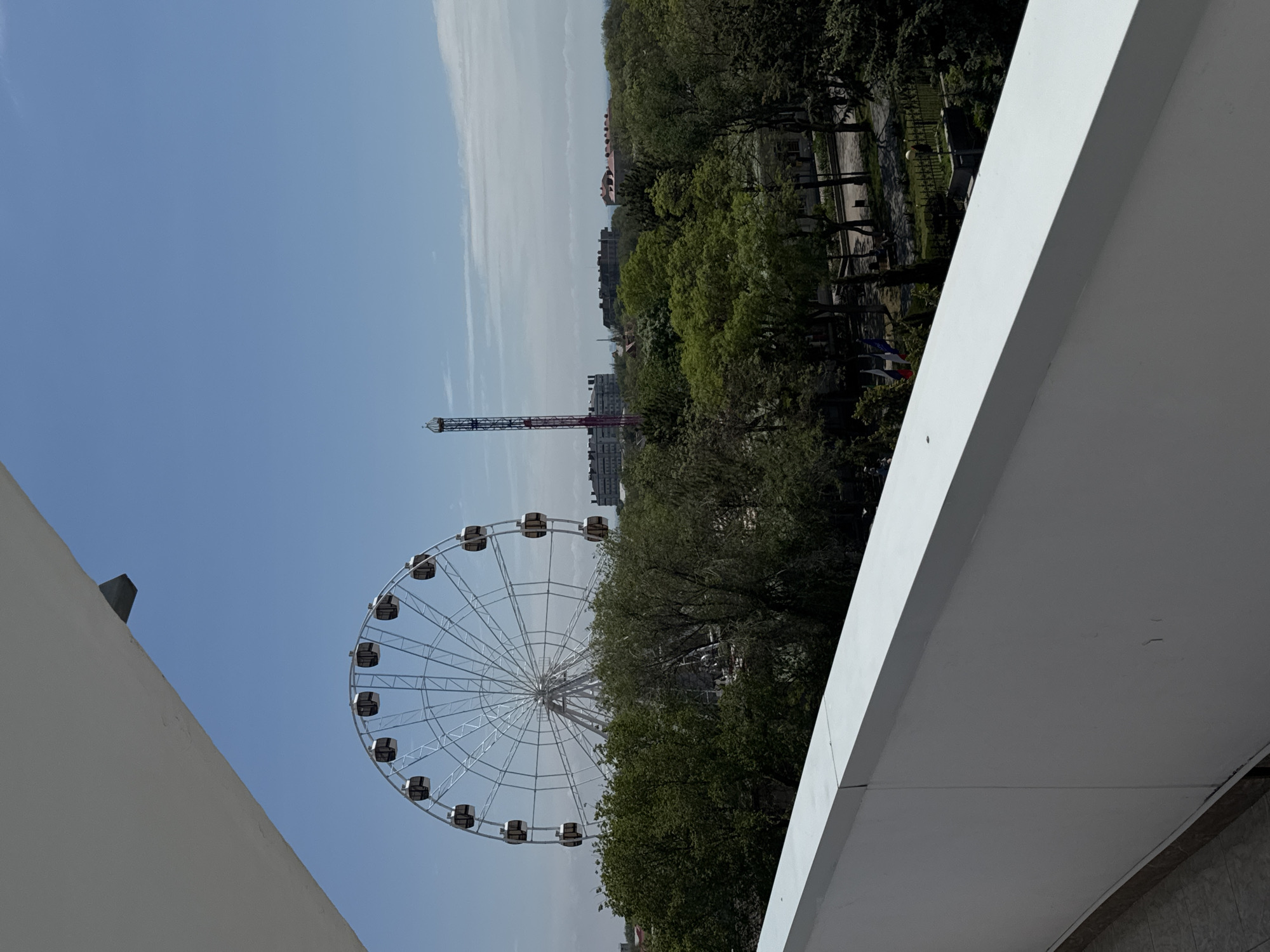 The image captures a partial view of Yevpatoriya, Ukraine, featuring a Ferris wheel and a tower amusement ride extending above a tree-filled park setting. Two white, modern architectural structures frame the shot on either side, creating a distinct foreground. The sky is predominantly blue with faint wisps of clouds, suggesting a clear day. Details such as trees, park infrastructure, and building outlines behind the park add depth to the urban panorama.