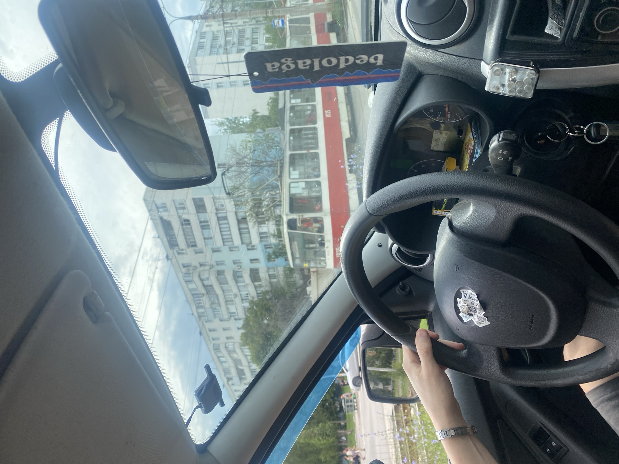 The image is taken from inside a car, looking forward through the windshield. A person's hands are on the steering wheel, indicating they are driving. Outside the car, a red and white tram is visible, along with apartment buildings and trees. The scene appears to be a street in Samara, Russia, given the context provided. A car air freshener with the brand name "bedolaga" is hanging from the rearview mirror.