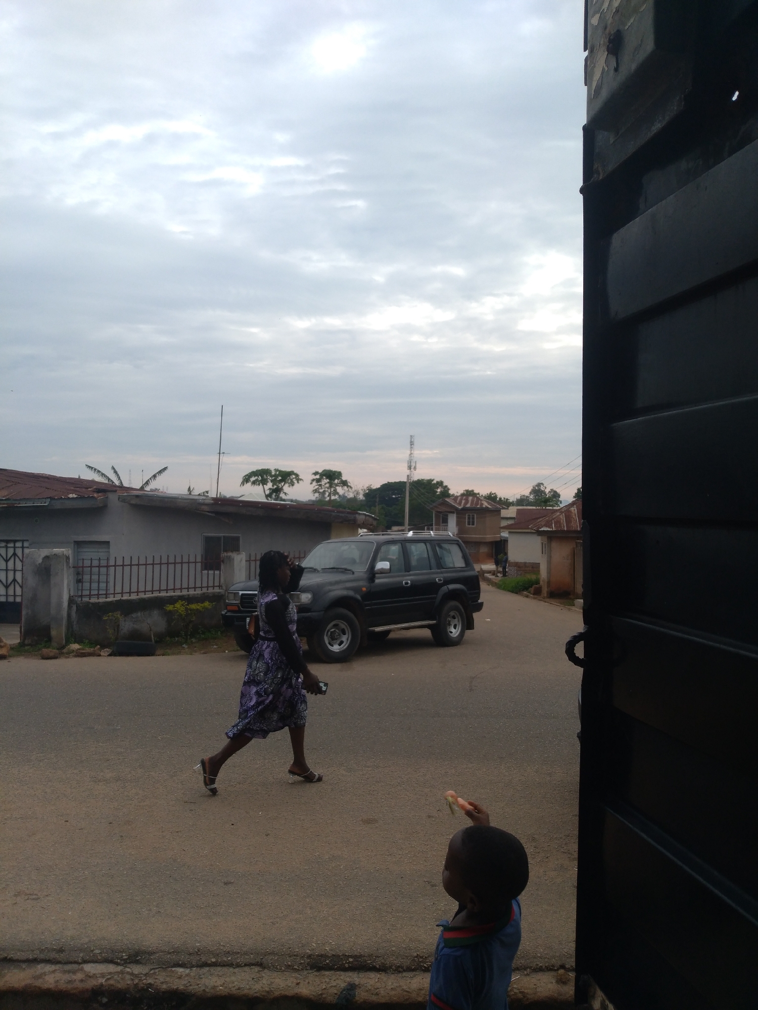 The scene depicts a street in Jos, Nigeria, under an overcast sky, suggesting diffused natural light. The foreground features an asphalt road with a concrete curb running horizontally across the bottom.
An adult female walks from left to right across the mid-ground, her body in profile facing right. She wears a long-sleeved, full-length dress patterned in purple and white, and open-toed sandals. Her dark hair appears tied back. She holds a light-colored mobile phone in her right hand, which is extended slightly forward.
Behind the walking woman, a dark-colored Toyota Land Cruiser SUV, appearing to be a J80 or J100 series, is positioned on the right side of the paved road, facing left. The vehicle has roof rails and alloy wheels.
In the background, residential buildings with corrugated metal roofs and painted or rendered walls line the street. On the left, a prominent single-story building painted gray features multiple security-barred windows and is fronted by a low concrete fence topped with metal railings. Further down the road, additional houses and a distant communication mast are visible amidst sparse tropical vegetation.
In the lower right foreground, a young child, likely a toddler, is partially visible, viewed from a slightly elevated perspective. The child wears a blue short-sleeved top with red and green trim on the collar. The child's head is tilted upwards and slightly to the left, and the right hand holds a reddish-pink slice of fruit, consistent with watermelon, which is held aloft.
The far right side of the image is vertically framed by a dark, partially open metal gate or doorframe, revealing a heavy-looking hinge or handle mechanism on its inner edge.