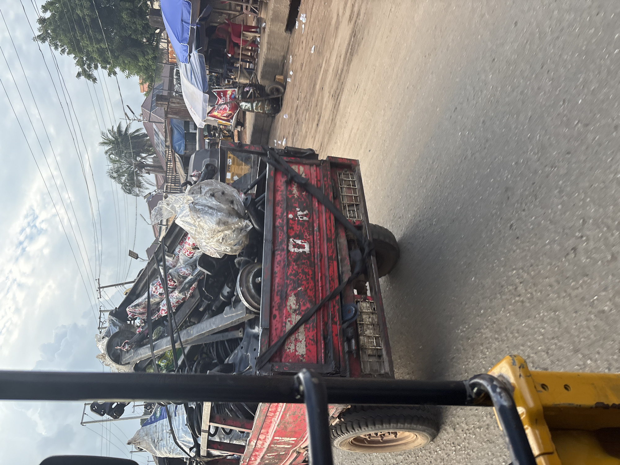 A red, three-wheeled vehicle, possibly a modified tuk-tuk or truck, is overloaded with metal parts and other items. The vehicle is moving on a paved road in Onitsha, Nigeria. The cargo is secured with straps and plastic wrapping. In the background, there are market stalls with blue and white awnings, trees, and utility poles. The sky is overcast with scattered clouds. The scene appears to be a busy street or market area.