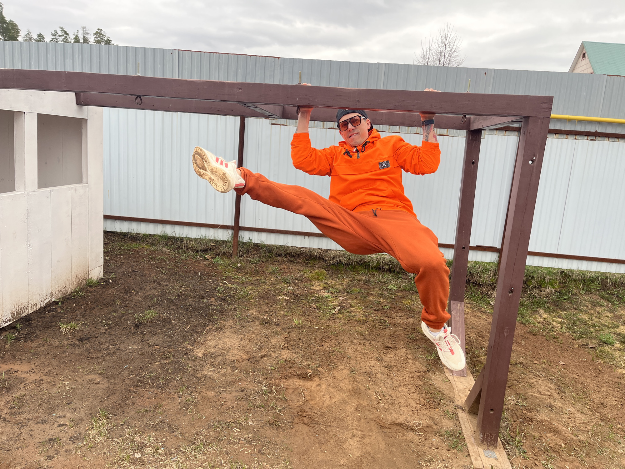 A man in an orange tracksuit performs an outdoor exercise in Chaikovsky, Russia. He is depicted in mid-air, holding onto a sturdy brown wooden pull-up bar with both hands. His right leg is extended horizontally outwards in a wide split, while his left leg is bent downwards. He wears a bright orange hooded jacket with a dark emblem on the chest, matching orange sweatpants, white sneakers with red stripes and muddy soles, a black baseball cap worn backward, and dark sunglasses. His expression is focused and determined as he looks towards the viewer.
The exercise setup is a simple wooden frame consisting of two vertical posts supporting the horizontal bar. To the left, part of a white, painted wooden structure or shed is visible. The background features a tall corrugated metal fence, behind which sparse trees and a distant building with a green roof can be seen under an overcast sky. The ground below is predominantly dirt and mud with patches of dry, sparse grass, indicating a potentially damp or unmaintained area. A yellow pipe runs along the top of the metal fence on the right side of the frame.
