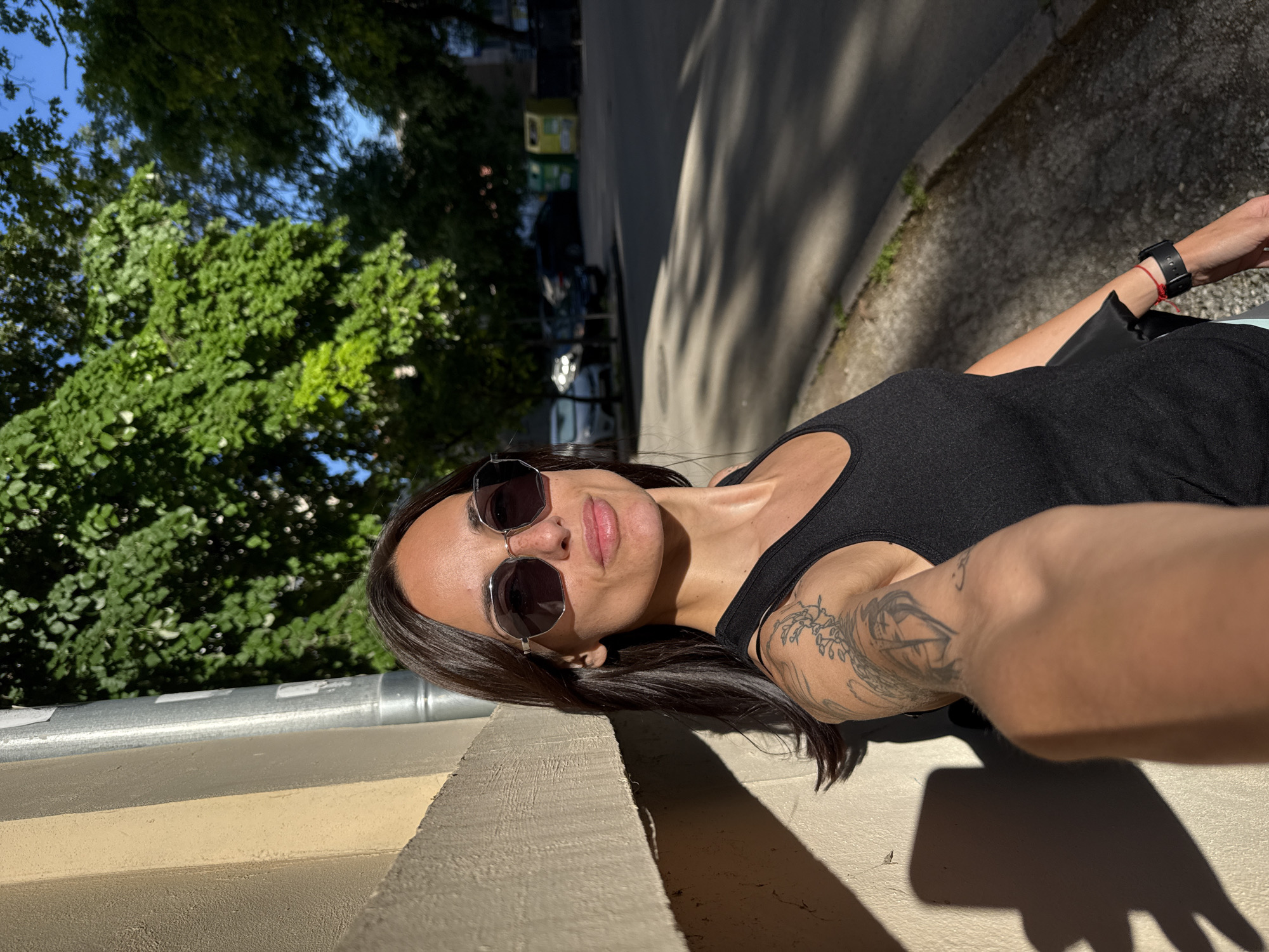 A woman wearing sunglasses and a black tank top is taking a selfie outdoors. She has dark hair and a tattoo on her left arm. She is leaning against a low wall with a metal pipe running along the top. Behind her, there are green trees, a road, and cars. The location is Pleven, Bulgaria.